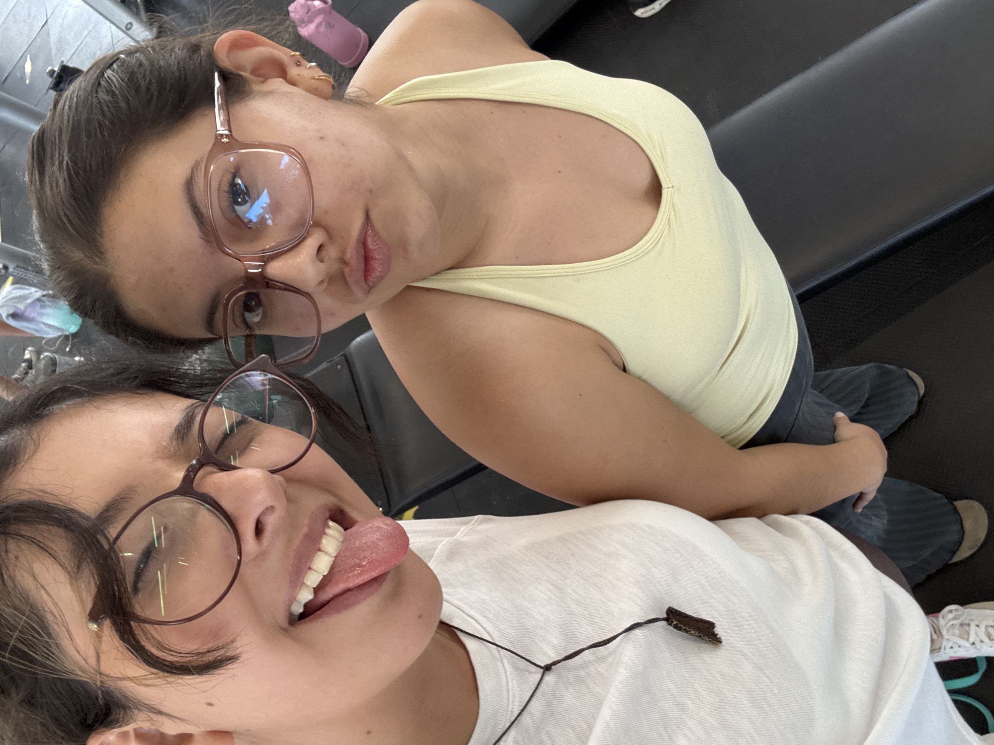 Two young women are posing for a selfie, likely indoors at a gym based on the equipment visible in the background. The woman on top is wearing a yellow tank top and glasses. The woman below is wearing a white t-shirt and glasses, sticking her tongue out at the camera. The location is San Andrés Cholula, Mexico.