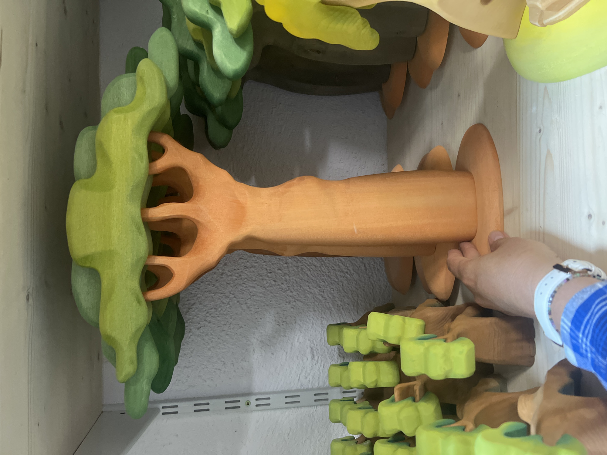 The image shows a close-up, overhead view of a collection of wooden toy trees. The trees are made of various colored wood, primarily orange, green, and yellow. They are designed in a way that suggests they are modular and can be assembled in different configurations. One of the trees, an orange one, has a trunk with a root-like base and what appears to be branches or extensions. A person's hand, wearing a blue and white plaid shirt and a white watch, is visible on the right side of the image, interacting with one of the wooden pieces, possibly assembling or holding it.
The background consists of a light-colored, textured wall and a wooden shelf with visible metal brackets. There is no discernible text in the image. The lighting suggests an indoor environment, possibly a playroom or a store displaying toys. The overall impression is one of creative play or an organized display of wooden toys. The focus is on the texture and form of the wooden toys, with the hand adding a human element to the scene.