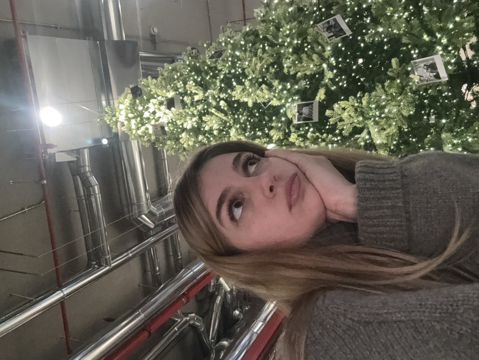 In a commercial or public indoor setting, city of Unknown, Unknown, a young adult female with light brown hair and a grey knitted sweater is depicted in a medium-close shot. She rests her right hand against her jawline, looking upward and to the right with a pensive expression. Behind her, dominating the right frame, stands a large, artificial Christmas tree adorned with numerous small white lights and several square photographic prints. The ceiling overhead features exposed silver and red industrial-style ventilation pipes and a bright, circular overhead light fixture.