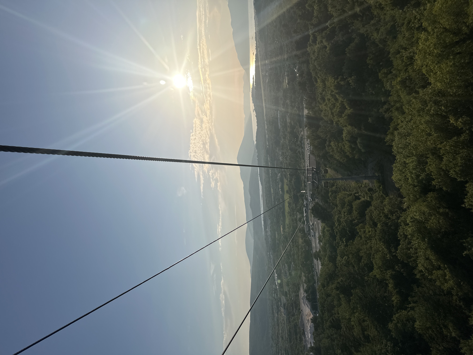 The image captures a view from a cable car ascending towards the summit of Mount Lovcen, overlooking the city of Dub, Montenegro. The sun, high in the sky and slightly to the right, casts a bright glare across the scene, its rays fanning out. Below, a lush green landscape stretches out, dotted with trees. In the middle ground, a body of water can be seen, reflecting the sunlight, with hills and mountains forming a dramatic backdrop. The cable car's thick wire runs diagonally across the frame from the upper left, a prominent element in the foreground. The overall impression is one of natural beauty and a panoramic vista.