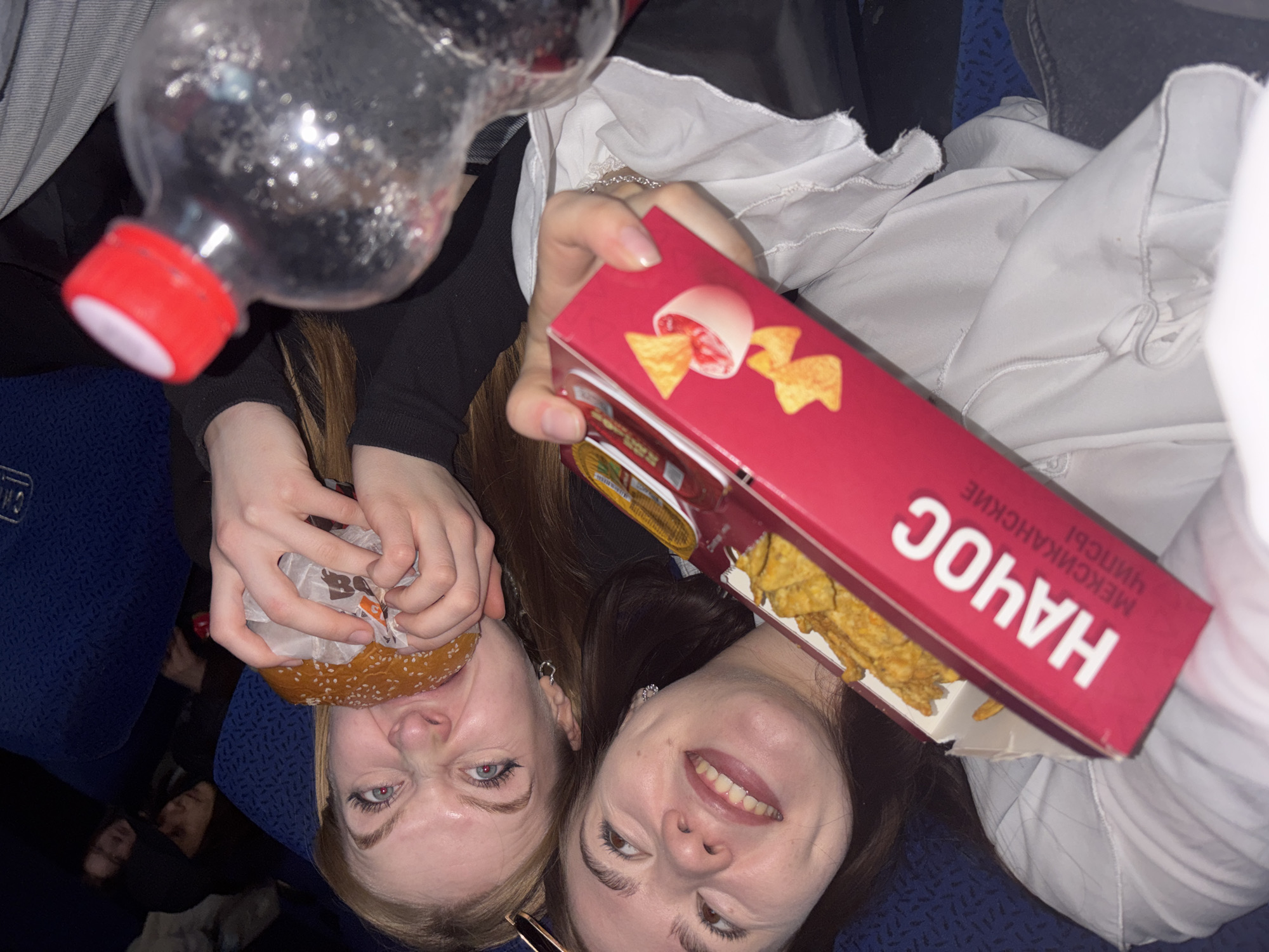 Two women are oriented upside down, facing the camera. The woman on the left is blonde and fair-skinned, holding a hamburger wrapped in paper with the Burger King logo. Her eyes are blue. The woman on the right is brunette and fair-skinned, holding a red box of "Nachos Mexicanos" chips, with some chips visible inside. Above them is a clear plastic bottle with a red cap, seemingly containing a dark liquid. They are seated on blue seating with a patterned design. Partially visible at the lower left are the faces of two more people. The environment suggests the seating of a public transport vehicle, possibly a bus or train. The location is Perm, Russia, based on the inscription "Мексиканские Чипсы" (Mexican Chips) on the nachos box, implying the image was taken in a Russian-speaking area.
