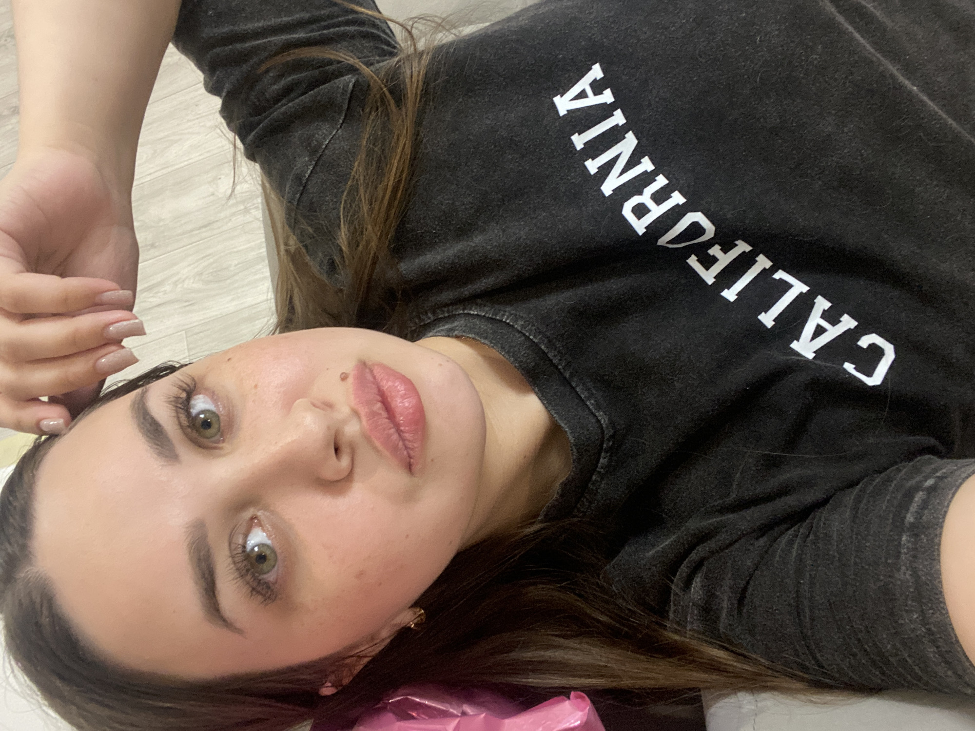 A selfie of a woman with brown hair, light skin, and green eyes wearing a black sweatshirt with the word "CALIFORNIA" printed on it in white, is taken in what appears to be an indoor setting. The background seems to be a light wood-like surface. There is a pink shiny object in the bottom corner of the picture. There is no indication of the location being in Chaikovsky, Russia.