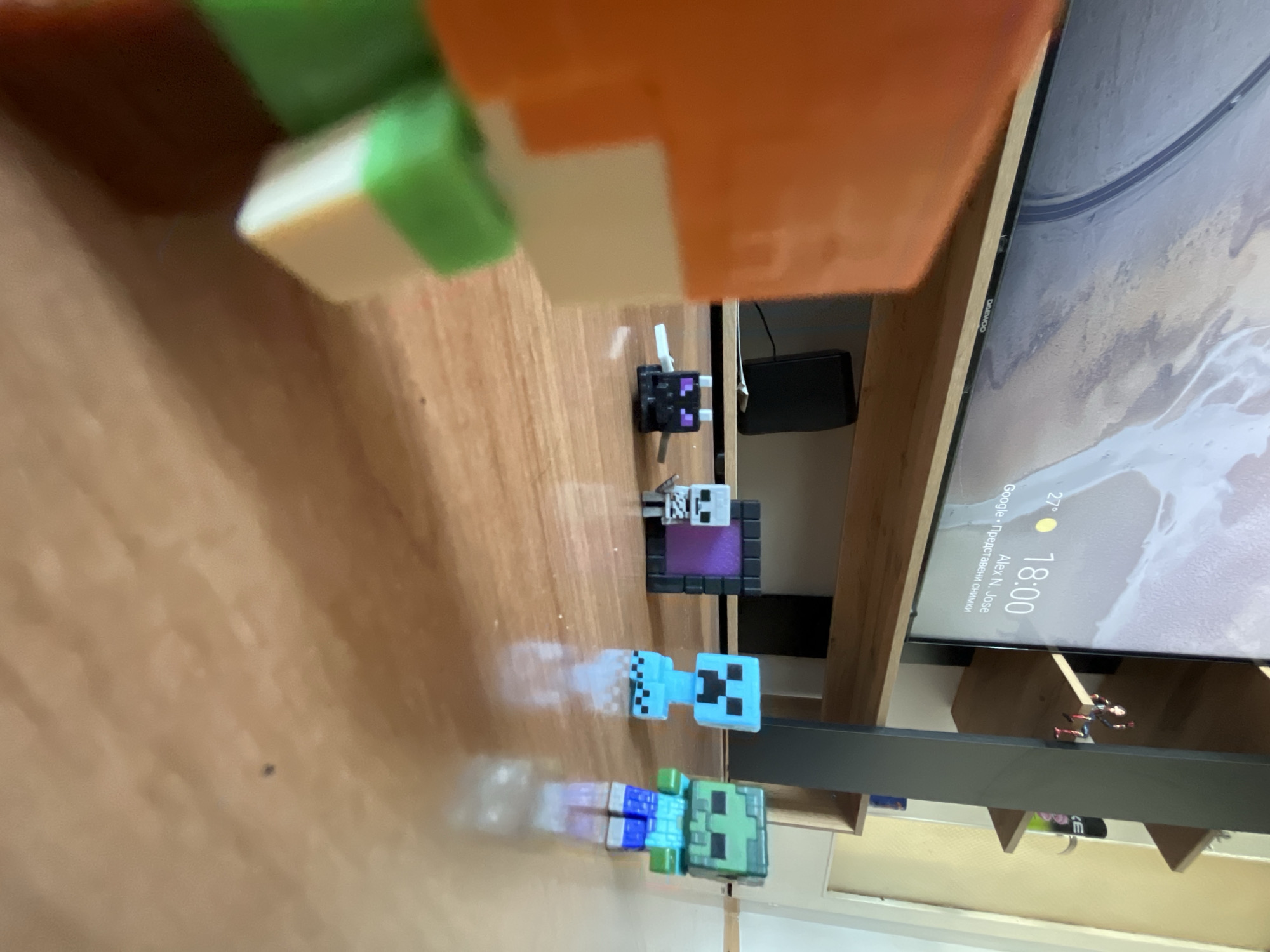 In a dimly lit room, a wooden floor stretches out with a television screen displaying a map and the time "18:00" in the background. A wooden shelf unit, partially visible, holds a few figurines. In the foreground, on the floor, are several small Minecraft-themed toys: an Enderman, a skeleton, a Nether portal, a Creeper, and a zombie. The toys appear to be arranged for a scene, with the Enderman and skeleton in the foreground, and the portal, Creeper, and zombie behind them. The overall scene is domestic, possibly a child's play area.