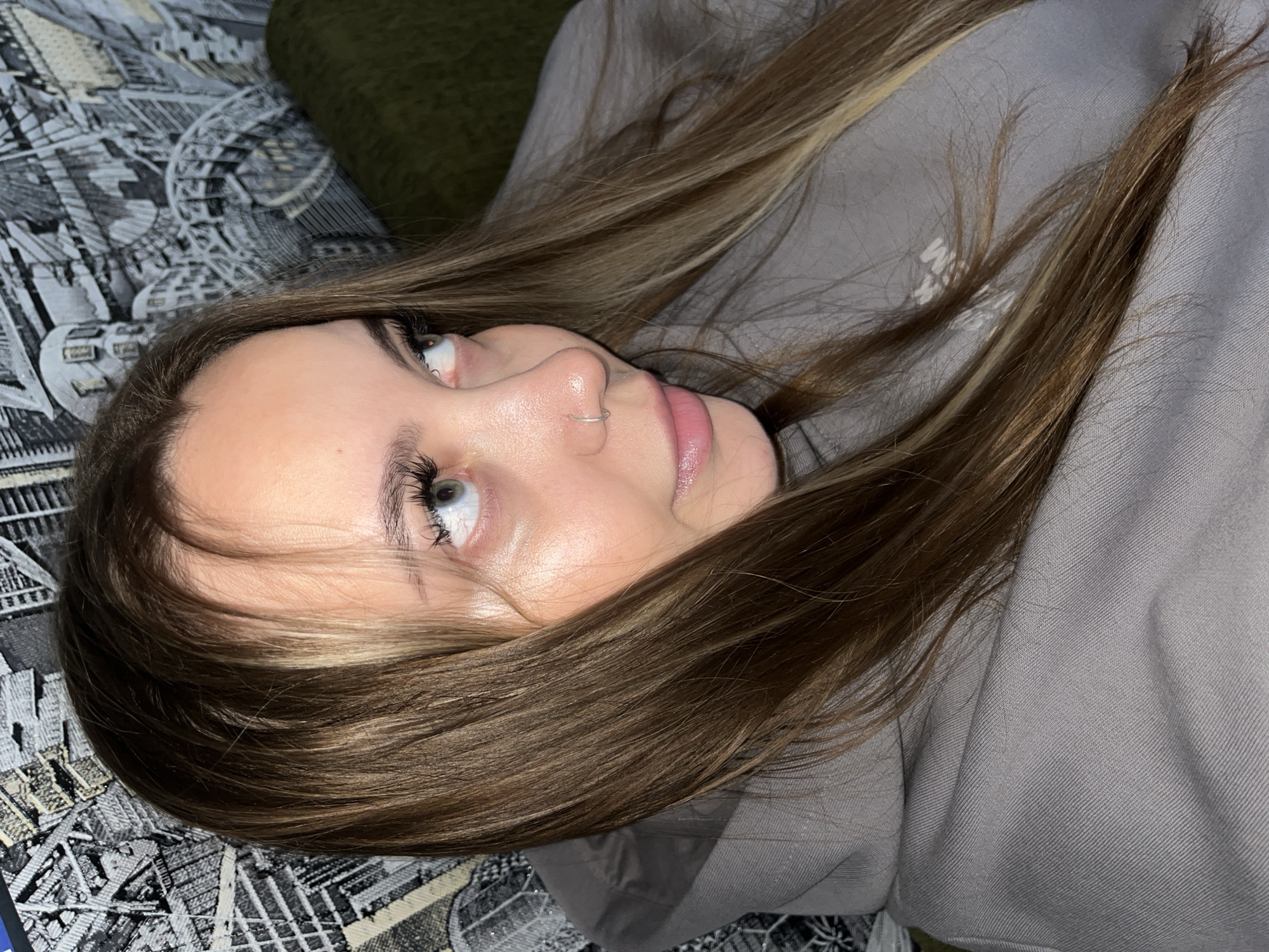 A young woman with long brown and blonde hair is lying on a patterned surface, wearing a gray hooded sweatshirt. She has a nose ring and is looking upwards to her left. The background includes a green object and the patterned surface on which she is lying. There is no indication of this image being taken in or near Novopokrovskaya, Russia.
