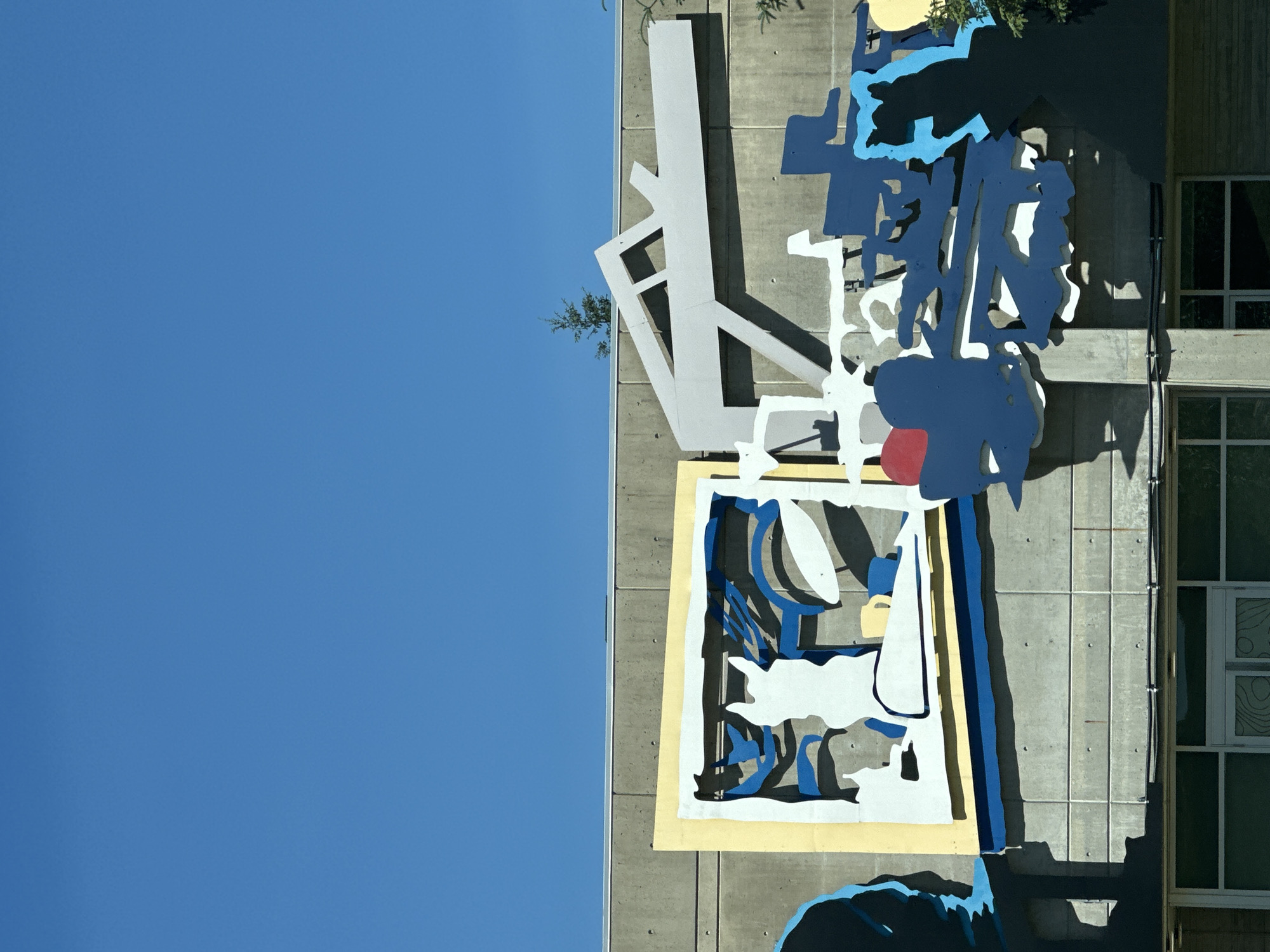 A section of a concrete building in Burnaby, Canada, features a colorful, abstract mural with geometric shapes and silhouetted figures against a clear blue sky. The mural is composed of flat, interlocking panels of various colors, including white, yellow, blue, light gray, and a small red circle.
One prominent element is a white silhouette of a dog or cat within a yellow-bordered frame, set against a background of blue and black abstract lines. To the right of this, a large, light gray angular structure extends horizontally, casting shadows. Below this, more blue and white abstract shapes are arranged, with a dark blue silhouette of a person or animal on the far right, partially obscured by a tree branch.
The building itself is constructed of grey concrete blocks. Below the mural, a row of windows and a doorway are visible. The overall scene suggests an outdoor installation on a sunny day.