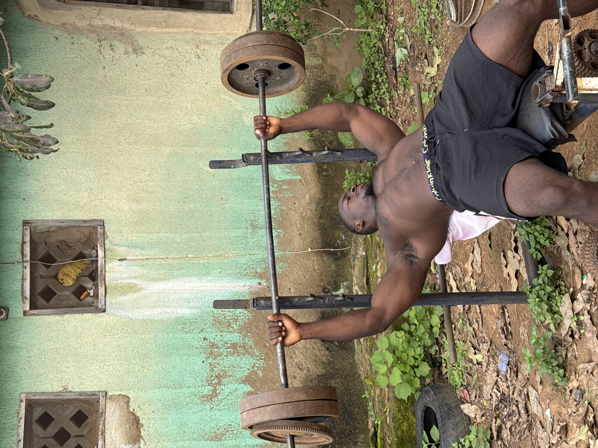 A Black man is performing a bench press outdoors in Akwanga, Nigeria. He is lying on a weight bench with a barbell loaded with rusty weight plates across his chest. The man is muscular, shirtless, and wearing black shorts with a Calvin Klein waistband. He is gripping the barbell with both hands, his eyes are closed, and his facial expression suggests exertion. The bench and barbell appear to be in a state of disrepair. Behind him is a weathered, light green wall with two square windows. The ground is covered with dirt, fallen leaves, and some sparse vegetation. A discarded tire is visible to the left of the bench. The overall environment suggests a somewhat neglected outdoor space.