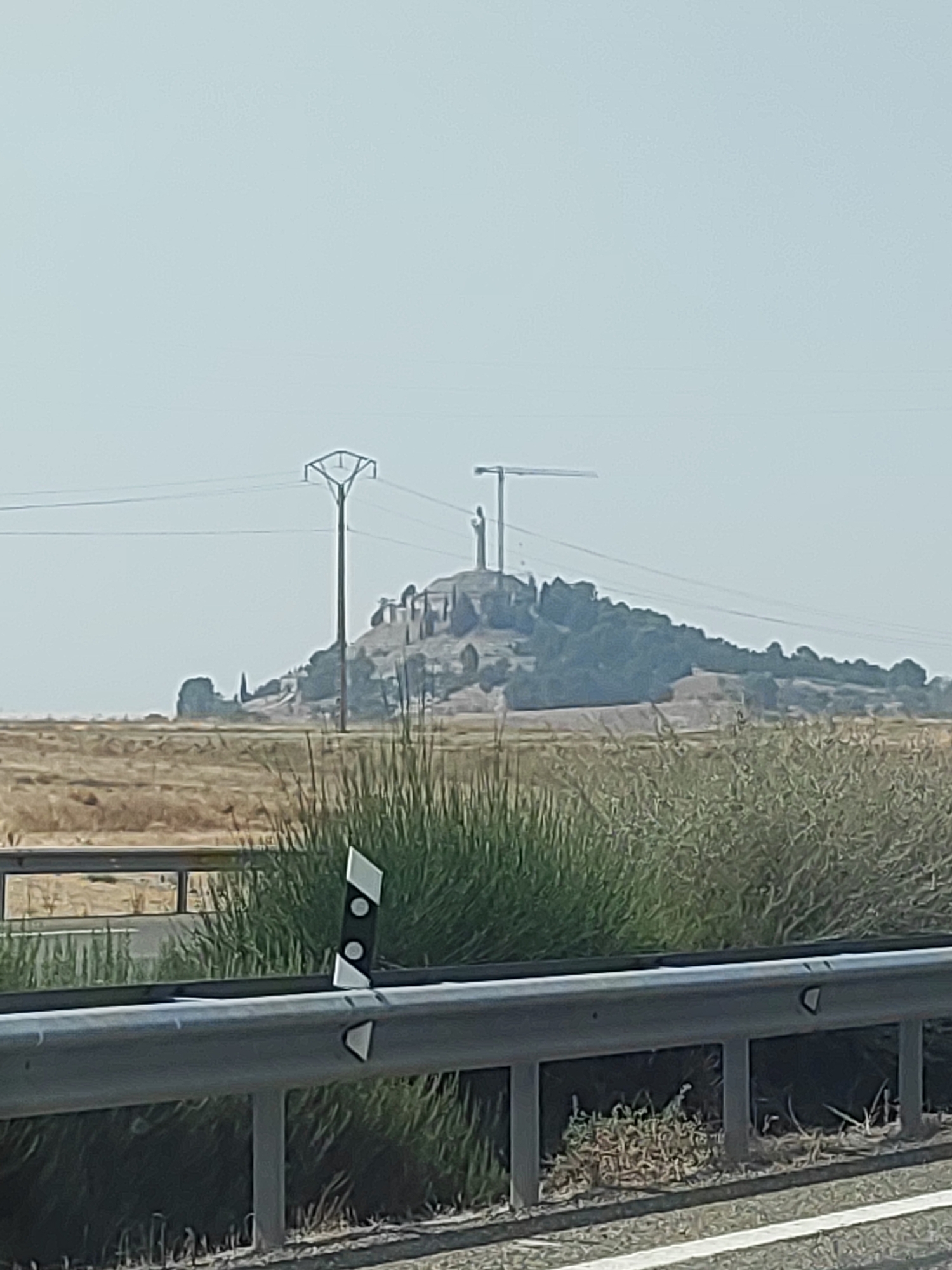 The image depicts a distant view of the Cristo del Otero monument in Palencia, Spain. The monument, which features a large statue of Christ on a hill, is partially obscured by foliage and appears to be under construction or renovation, as indicated by the presence of a crane. The scene is set against a clear sky and a dry, arid landscape. In the foreground, a metal guardrail separates the viewer from a grassy area and a road. The overall impression is one of a vast, open space with a prominent landmark in the distance.