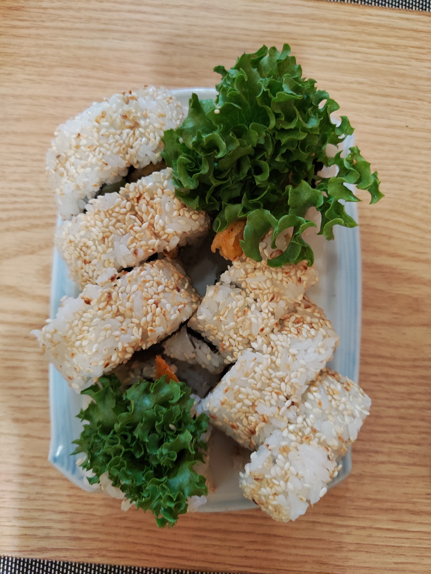 A top-down shot captures a rectangular ceramic plate with a light blue border, holding sesame-covered sushi rolls and green lettuce. The sushi pieces are arranged somewhat haphazardly, and the lettuce seems to be used as a garnish. The plate rests on a light brown wooden surface with a patterned border visible at the bottom of the image. The scene depicts a simple meal setting. The city of Da Lat, Vietnam, is not directly visible.