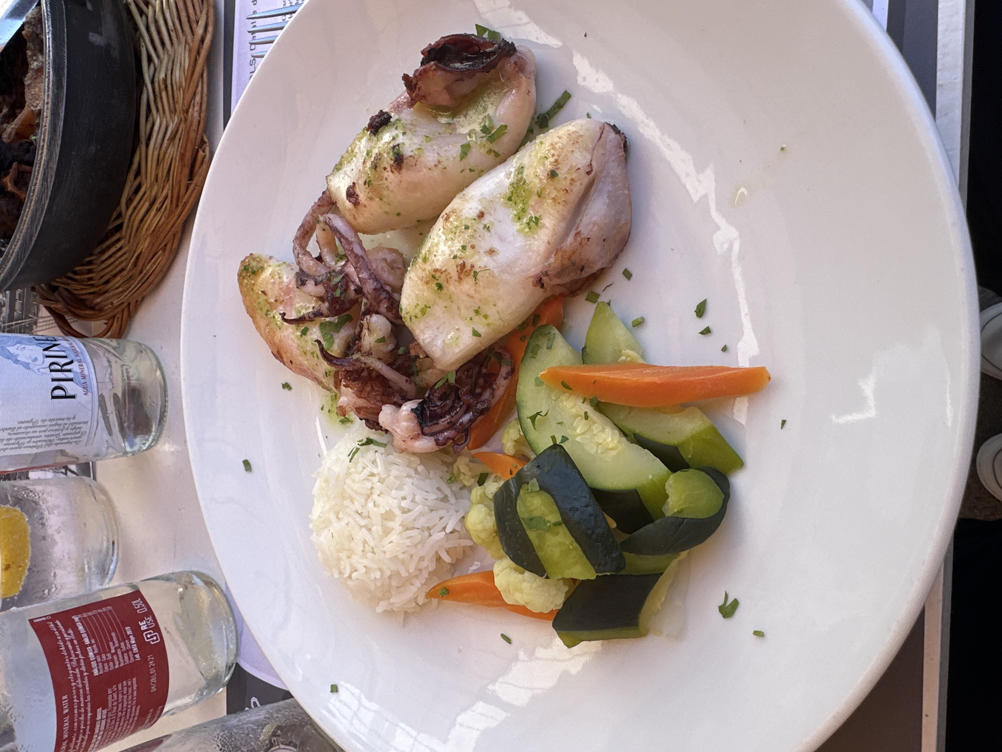 A top-down, close-up shot reveals a white plate piled with food, presumably in Sant Feliu de Guíxols, Spain. The main component of the dish is what appears to be grilled squid, seasoned with herbs and charred in places. Alongside the squid are portions of white rice and cooked vegetables, including sliced cucumbers, carrot sticks, and pieces of cauliflower. The plate rests on a table surface next to several glass bottles of mineral water, labeled "PIRINE." A wicker basket containing an unidentifiable dish is partially visible on the left side of the frame.