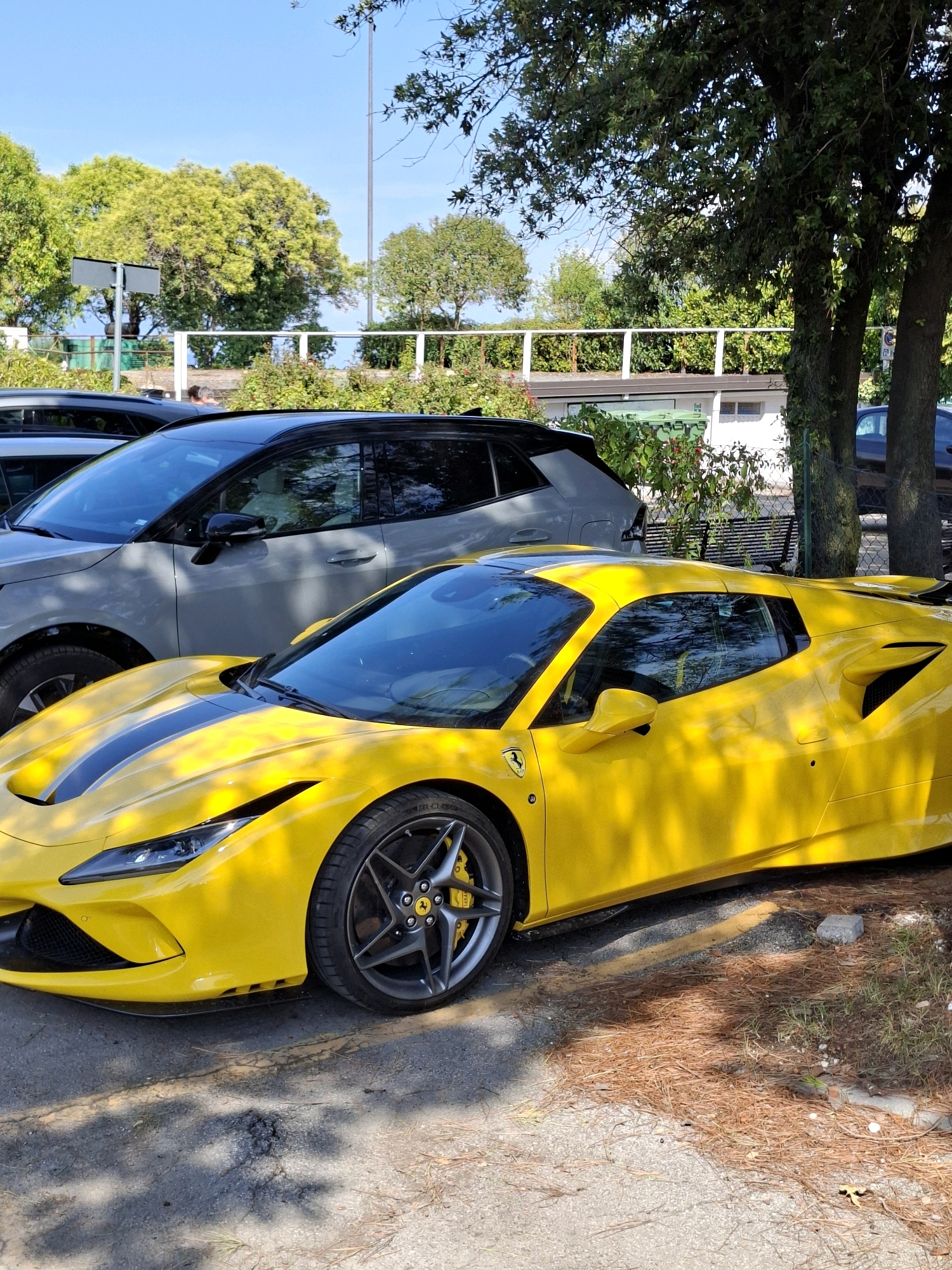A bright yellow Ferrari F8 Tributo is parked in what appears to be a parking lot in Sirmione, Italy. The car is positioned in the foreground, with its sleek lines and distinctive racing stripes clearly visible. Behind the Ferrari, a light gray SUV is parked, suggesting a shared parking area. The environment includes trees casting dappled shadows on the ground, indicating a sunny day. In the background, there are hints of buildings and vegetation, typical of a Mediterranean setting. The overall scene conveys a sense of luxury and leisure, possibly a vacation spot.