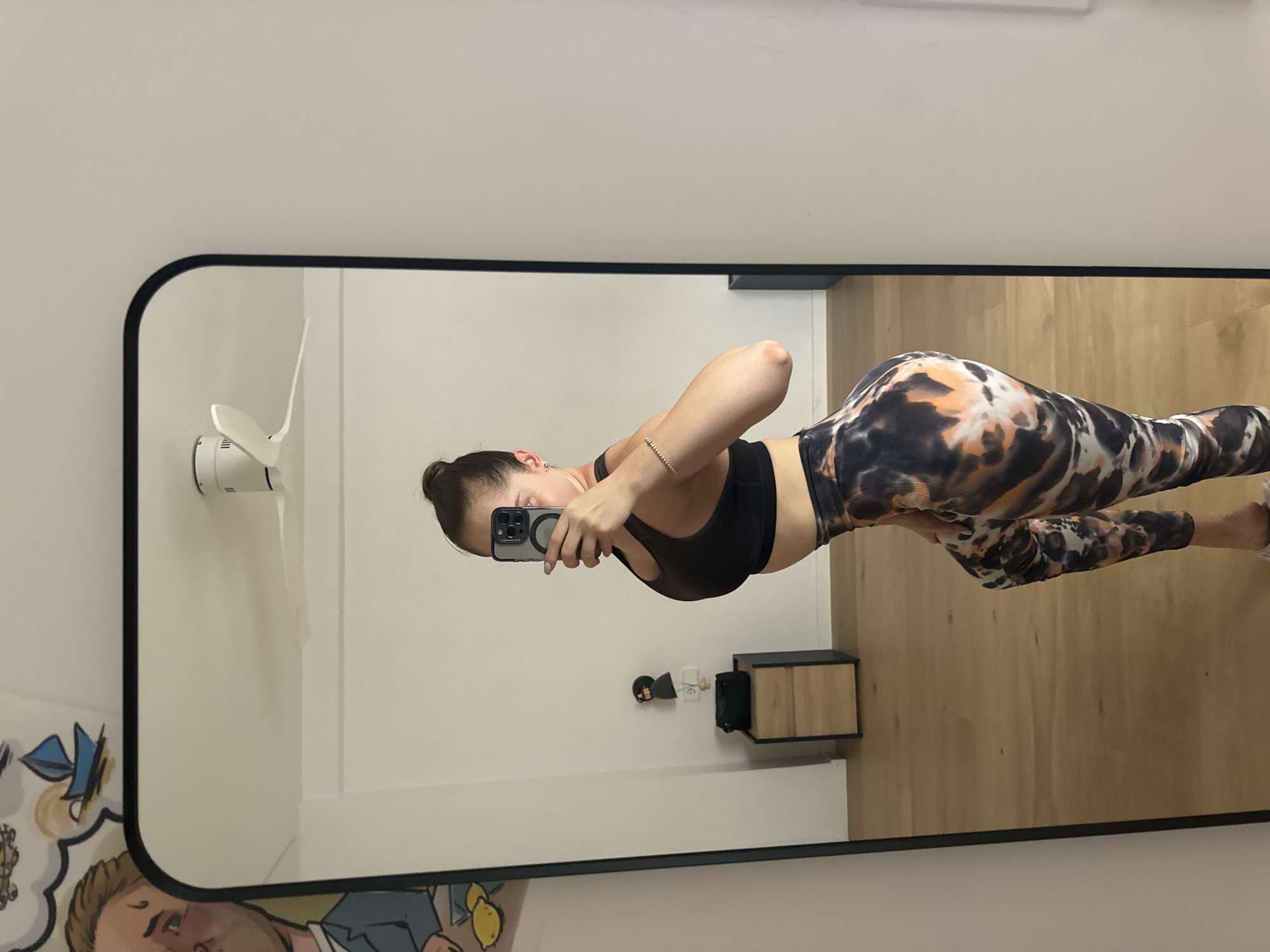 A full-length mirror reflects a woman in athletic wear. She is wearing a black sports bra and leggings with a black, white, and orange tie-dye pattern. The woman is holding a smartphone in her right hand, taking a selfie. Her hair is pulled back in a bun. In the reflection of the mirror, a white ceiling fan is visible on the left, and a light-colored wall. A small black cabinet with wooden drawers is on the right. Below the mirror, part of a colorful mural is visible. The setting appears to be an indoor space, likely a home or gym. There is no indication of the time of day or weather. No visible text is present in the image. The image is oriented vertically.