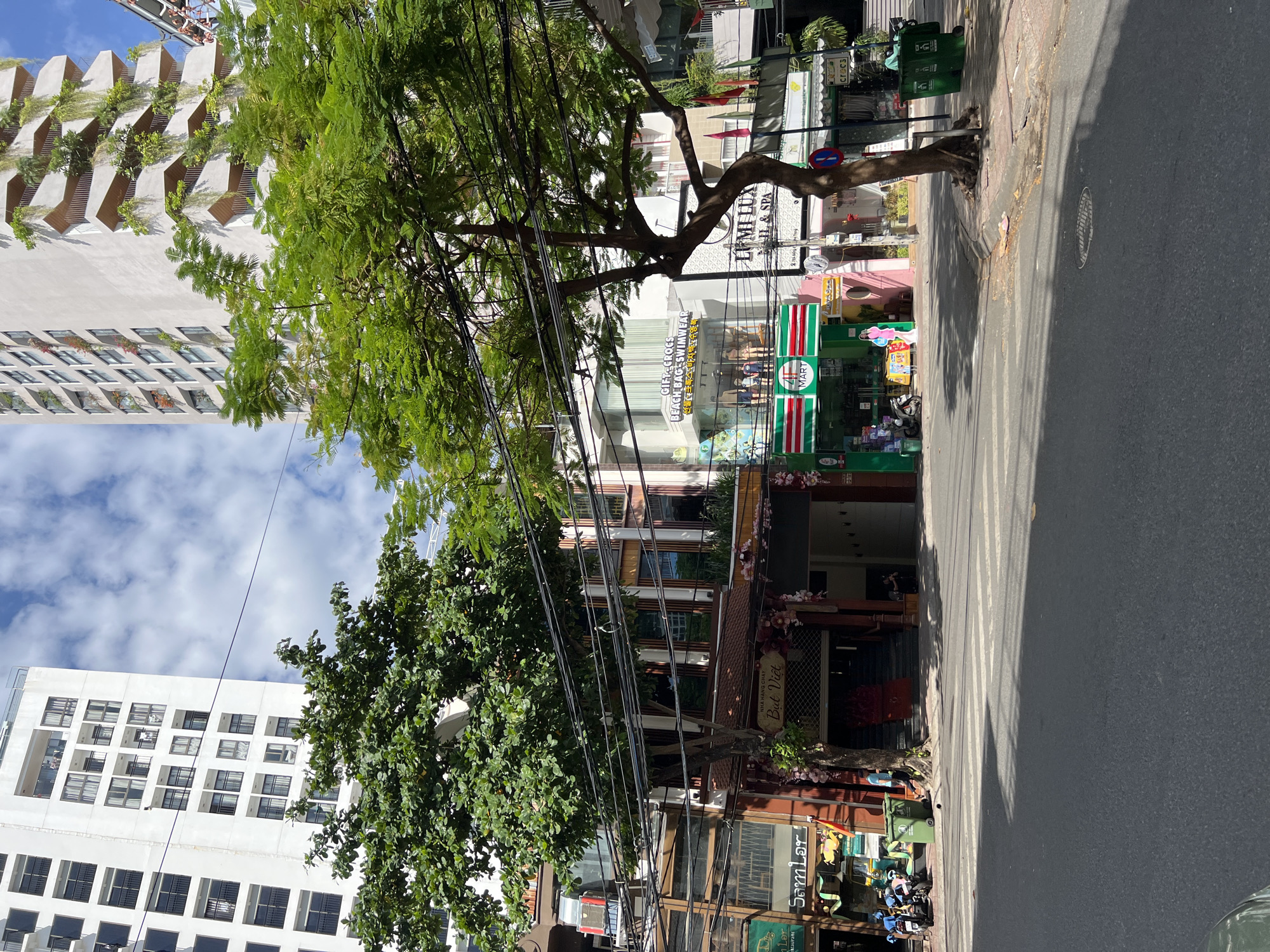 In Nha Trang, Vietnam, a street scene unfolds under a partly cloudy sky. Two tall buildings, one stark white with repeating window patterns and another with a more organic, tiered design featuring abundant greenery, dominate the background. In the middle ground, a street lined with shops and businesses runs horizontally. The shops have awnings and display windows, with some signs visible in Vietnamese. A prominent tree with lush green foliage stands in front of the buildings, its branches reaching over the street and obscuring some of the architecture. Overhead, a tangle of electrical wires crisscrosses the sky. The foreground shows a paved street with a crosswalk, and on the right side of the street, green trash receptacles are visible. A few parked motorcycles and a person walking are also present.