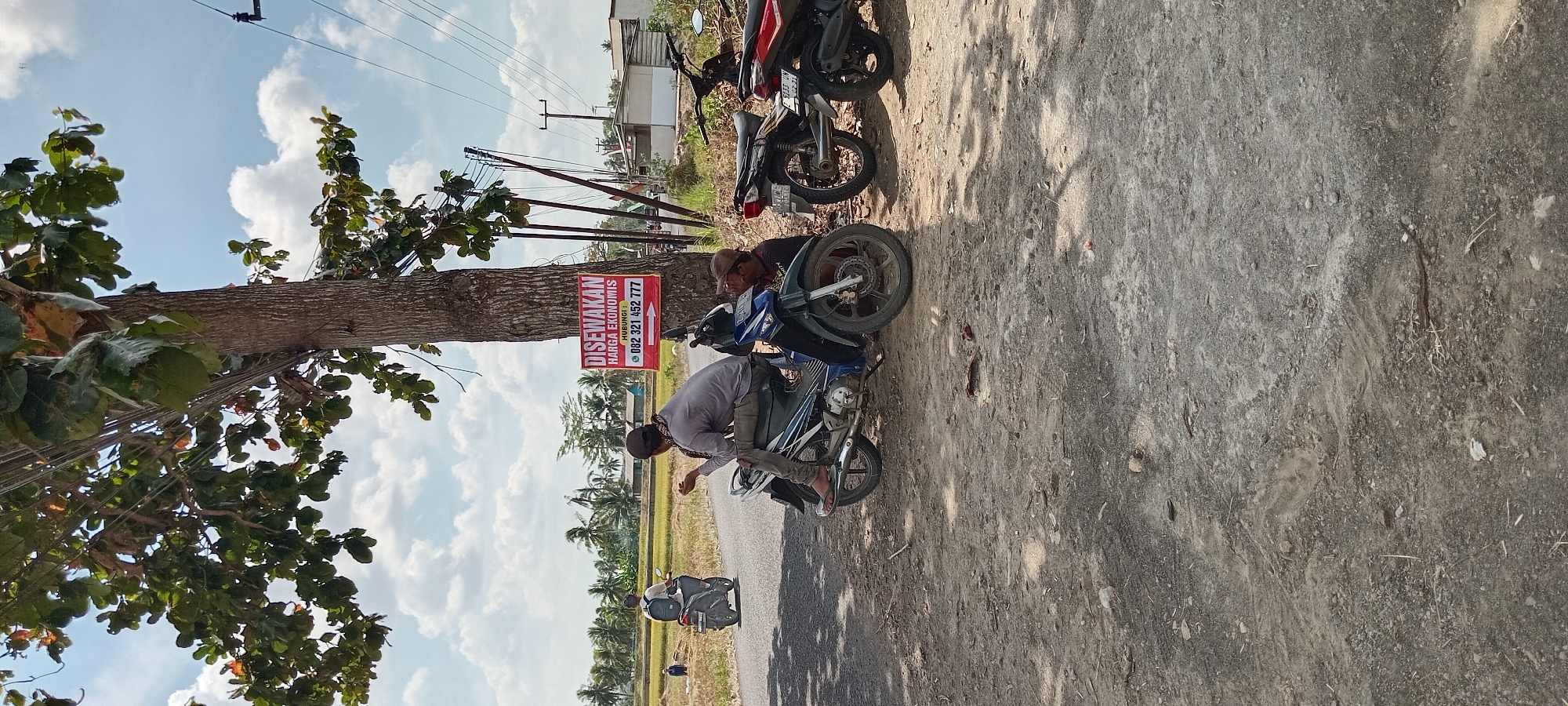 In Karangpawitan, Indonesia, on a sunny day with scattered clouds, a street scene unfolds near a rural landscape. A prominent tree with a thick trunk stands on the right side of the frame, its branches reaching towards the sky, partially obscuring power lines. Attached to the tree is a red sign advertising "DISEWAKAN" (for rent) with the text "HARGA EKONOMIS" (economical price) and a phone number, "082 321 452 777".
In the foreground, two men are casually seated on a blue motorcycle parked on the side of a paved road. One man, wearing a gray long-sleeved shirt and dark pants, is looking down, possibly at his phone or something on the ground. The other man, wearing a black shirt and brown cap, is partially visible, leaning against the motorcycle. Further down the road, another motorcycle, black and red, is parked.
In the background, a motorcycle with a rider is moving away from the camera on the road. Beyond the road, a vast expanse of rice paddies stretches out, with a few scattered houses and coconut trees dotting the horizon. In the distance, a person can be seen working in the rice paddy. The ground in the foreground is a mix of dirt and gravel, with some dried leaves and debris scattered around. The overall impression is a quiet, rural setting with a hint of commercial activity indicated by the rental sign.