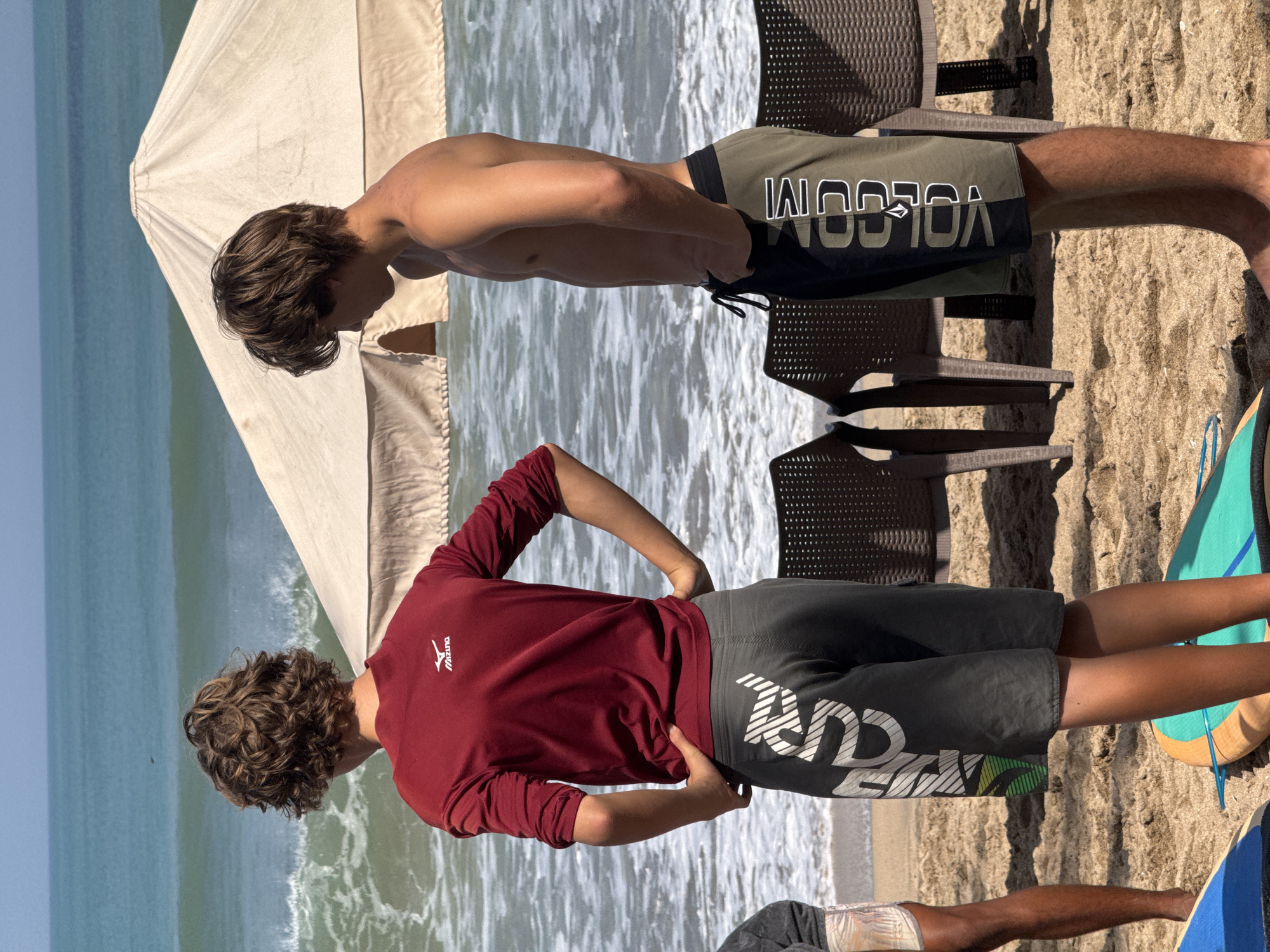 Two young males are standing on a sandy beach with the ocean and waves in the background. The male on the left wears a long-sleeved maroon shirt with a white logo on the back and dark grey shorts with white and green graphic designs. The male on the right is shirtless and wears olive green and black board shorts with white lettering. They are both facing away from the camera and looking out at the ocean. In the background, there is a large, light-colored umbrella and several brown chairs. The sea is a vibrant blue-green, and waves are breaking on the shore. Several surfboards are visible on the sand in the foreground. The scene appears to be a sunny day in Kuta, Indonesia.