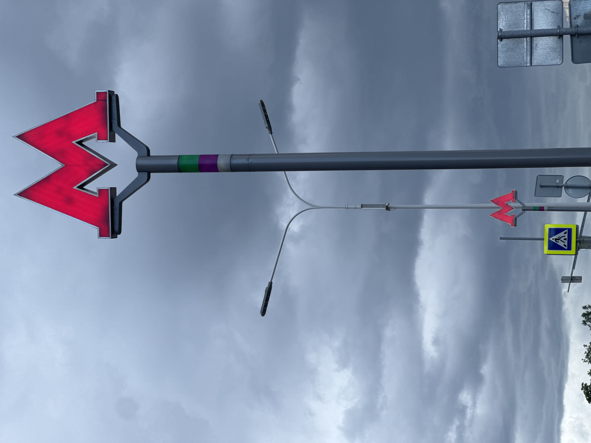 A street scene in Moscow, Russia, featuring a red "M" subway sign, a pedestrian crossing sign, and streetlights against a cloudy sky. The subway sign is prominently displayed on a metal pole with green and purple markings.
