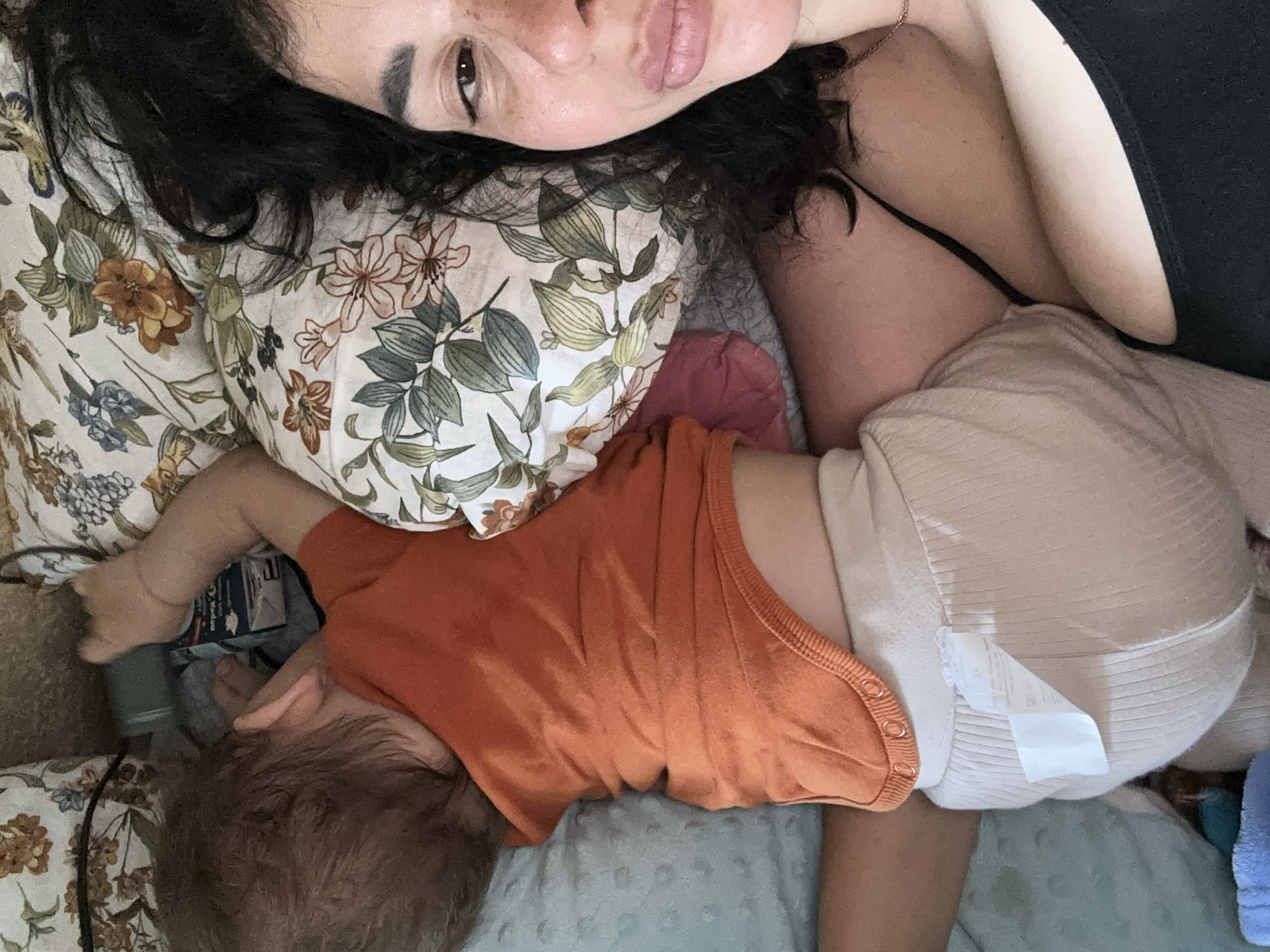 The image depicts a top-down, close-up view of a woman and a baby lying in bed. The baby is sleeping on its stomach, wearing an orange bodysuit and beige ribbed pants. The woman is lying beside the baby, with her head resting on a floral-patterned pillow. Her face is partially visible, and she appears to be looking at the camera. The environment suggests an indoor setting, likely a bedroom, with bedding and pillows visible. The overall scene conveys a sense of intimacy and rest.