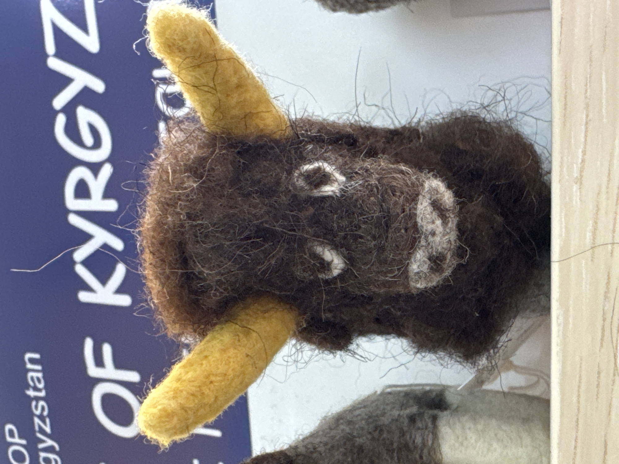 The image displays a close-up of a handcrafted, felted animal head, likely a yak or bull, positioned vertically. The animal's head is primarily dark brown, characterized by a shaggy and coarse texture composed of distinct fibers. It features two prominent, upward-curving yellow horns. Simple white circular markings serve as eyes, and a larger white, textured area forms the snout or muzzle.
The felted head is set against a vivid blue background, which appears to be part of a sign or informational display. White capital letters are clearly visible on this blue surface, spelling "OF KYRGYZ." Further down, partially visible text includes "OP" and "gyzystan," strongly suggesting the complete phrase is "SHOP OF KYRGYZSTAN." To the right of the animal head, a light-colored wooden or laminated surface is visible, possibly part of a display shelf or frame. A section of another felted object, featuring grey and white tones, is partially visible at the bottom of the frame. The image captures the fibrous detail of the felted material, emphasizing its artisanal quality. The visible text and the provided location context of Кашат, Kyrgyzstan, indicate that the item is a local craft or souvenir.
