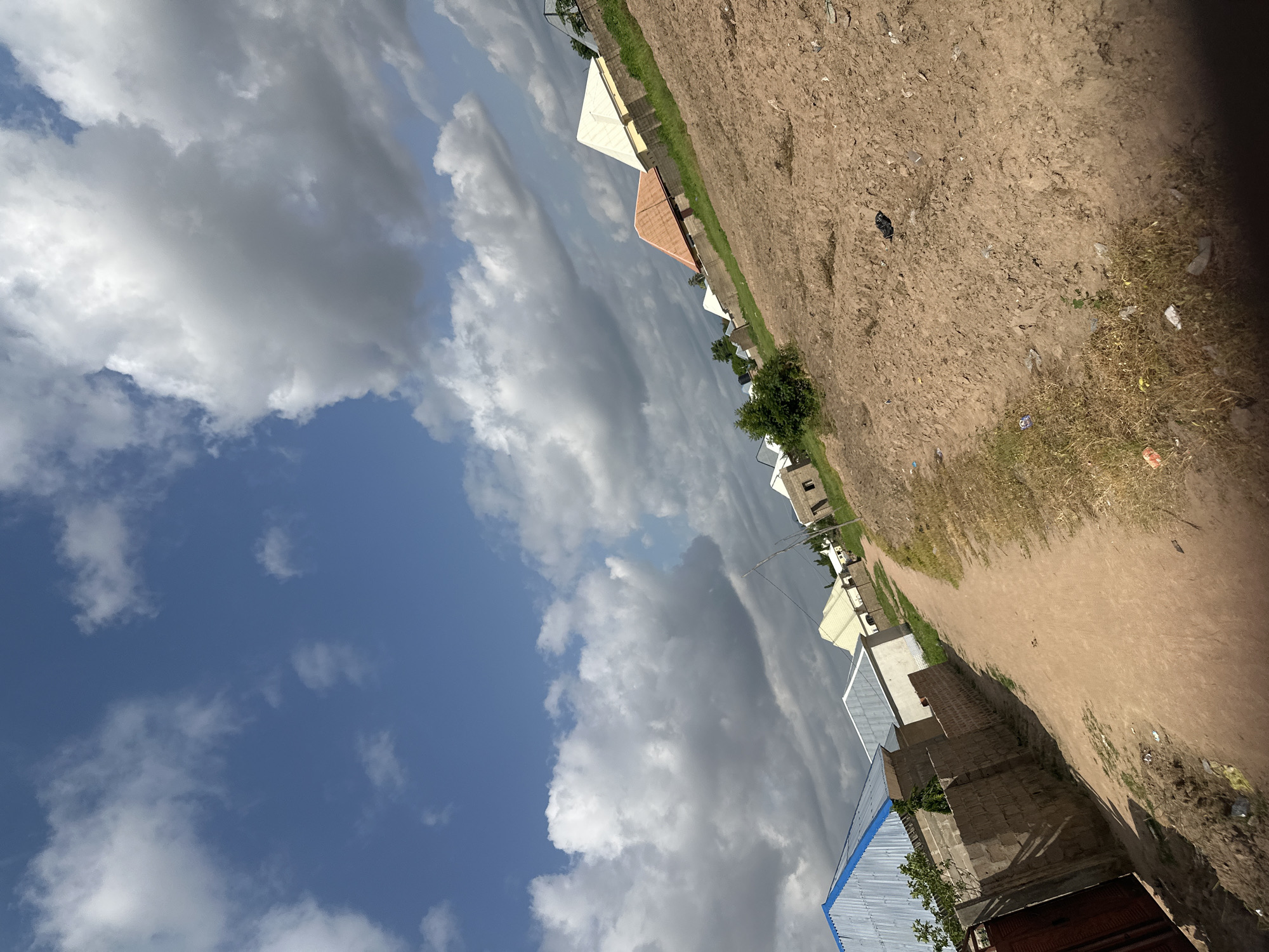 The image displays a street scene in Jalingo, Nigeria, characterized by a mix of residential buildings and an unpaved dirt road. The architecture suggests a developing area, with some structures featuring corrugated metal roofs and others appearing to be under construction with exposed brickwork. Scattered debris and patches of dry grass indicate a somewhat unkempt environment.
Dominating the left side of the frame is a dynamic sky filled with large, dramatic clouds. The blue sky peeks through in patches, suggesting a day with variable weather. The clouds range from bright white and puffy to dark and ominous, hinting at the possibility of rain or recent showers.
The perspective of the image is tilted, with the ground running diagonally from the bottom right to the top left. This creates a sense of depth and emphasizes the expanse of the sky. The sun appears to be casting shadows, suggesting it is likely daytime, possibly late morning or early afternoon given the light and cloud cover.
There is no visible text in the image. The overall impression is one of a rural or semi-urban setting in a tropical or subtropical climate, with a focus on the contrast between the natural elements of the sky and the man-made structures.