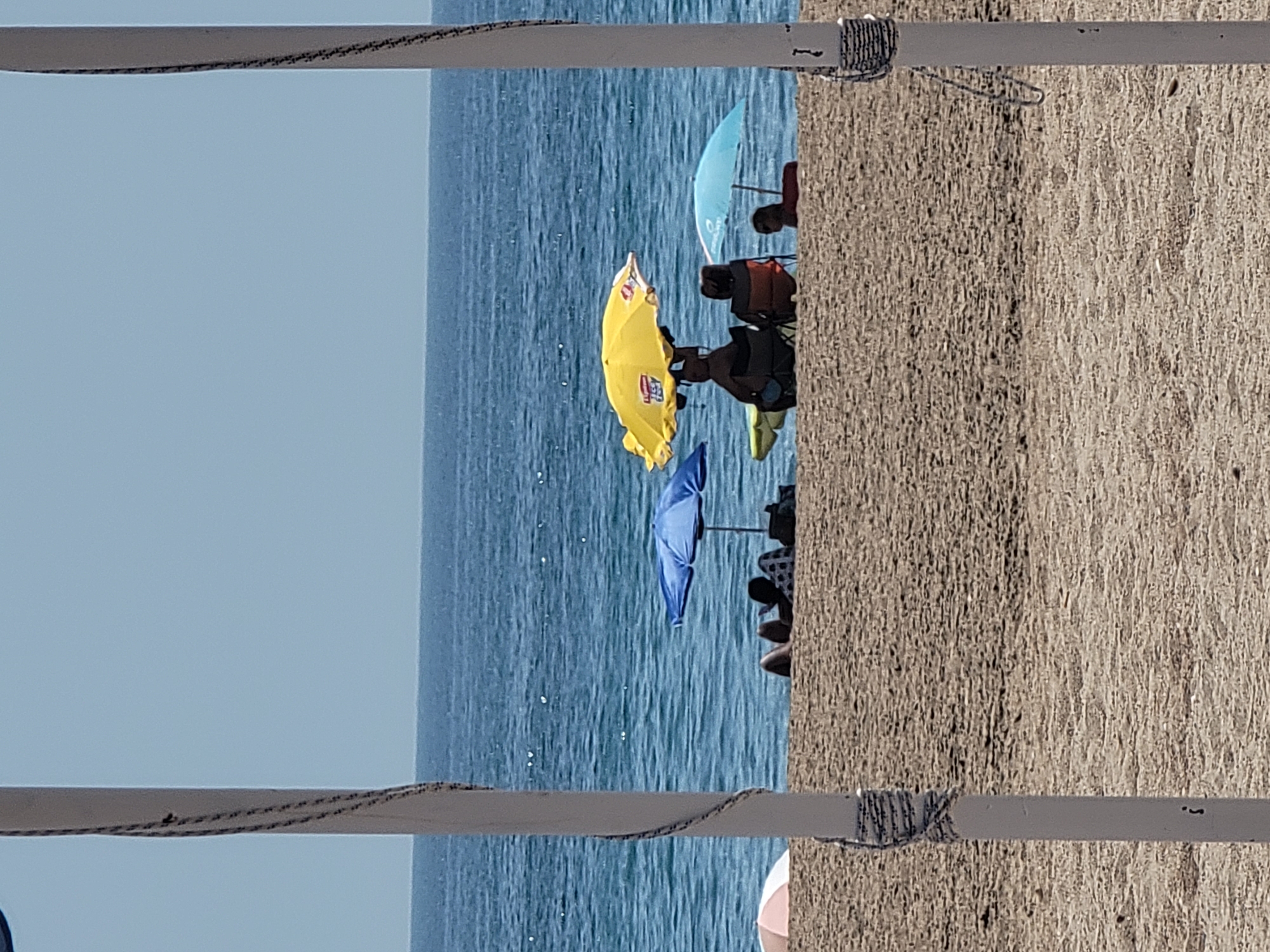 The image depicts a sunlit beach scene in Quarteira, Portugal. The foreground is dominated by the sandy texture of the beach, with two white poles, wrapped with rope, framing the view. In the mid-ground, several beachgoers are visible. A person sits under a blue umbrella reading, with another person next to them. Further to the right, a bright yellow umbrella shades individuals in beach chairs, and a light blue umbrella is also present. Beyond the beach, the tranquil blue ocean stretches to the horizon, meeting a clear, pale blue sky. The overall atmosphere is one of a peaceful summer day by the sea.