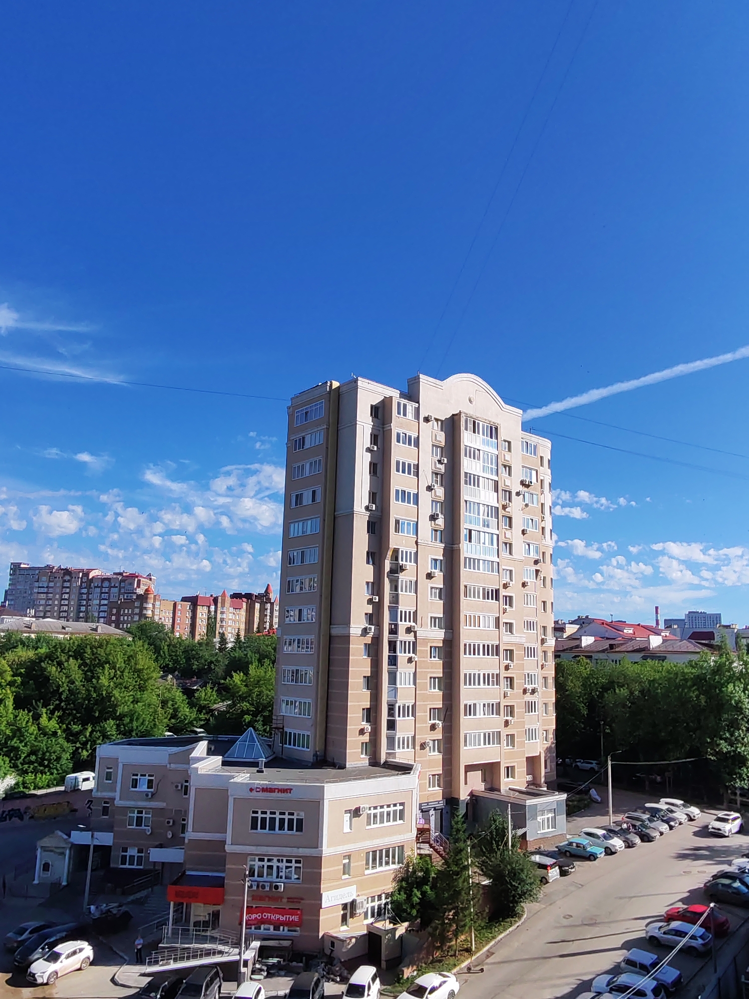 This image captures an urban street scene in Ufa, Russia, under a bright blue sky with scattered white clouds. Dominating the foreground is a tall, beige apartment building with numerous windows and air conditioning units. To the left of the apartment building is a lower, light-colored commercial building with a prominent red and white sign that reads "МАГНИТ" (Magnit), a Russian supermarket chain. Several cars are parked along the street and in a lot. In the background, other apartment buildings and trees are visible, indicating a densely populated residential area. The overall impression is a typical day in a Russian city, with modern architecture and everyday urban activity.
