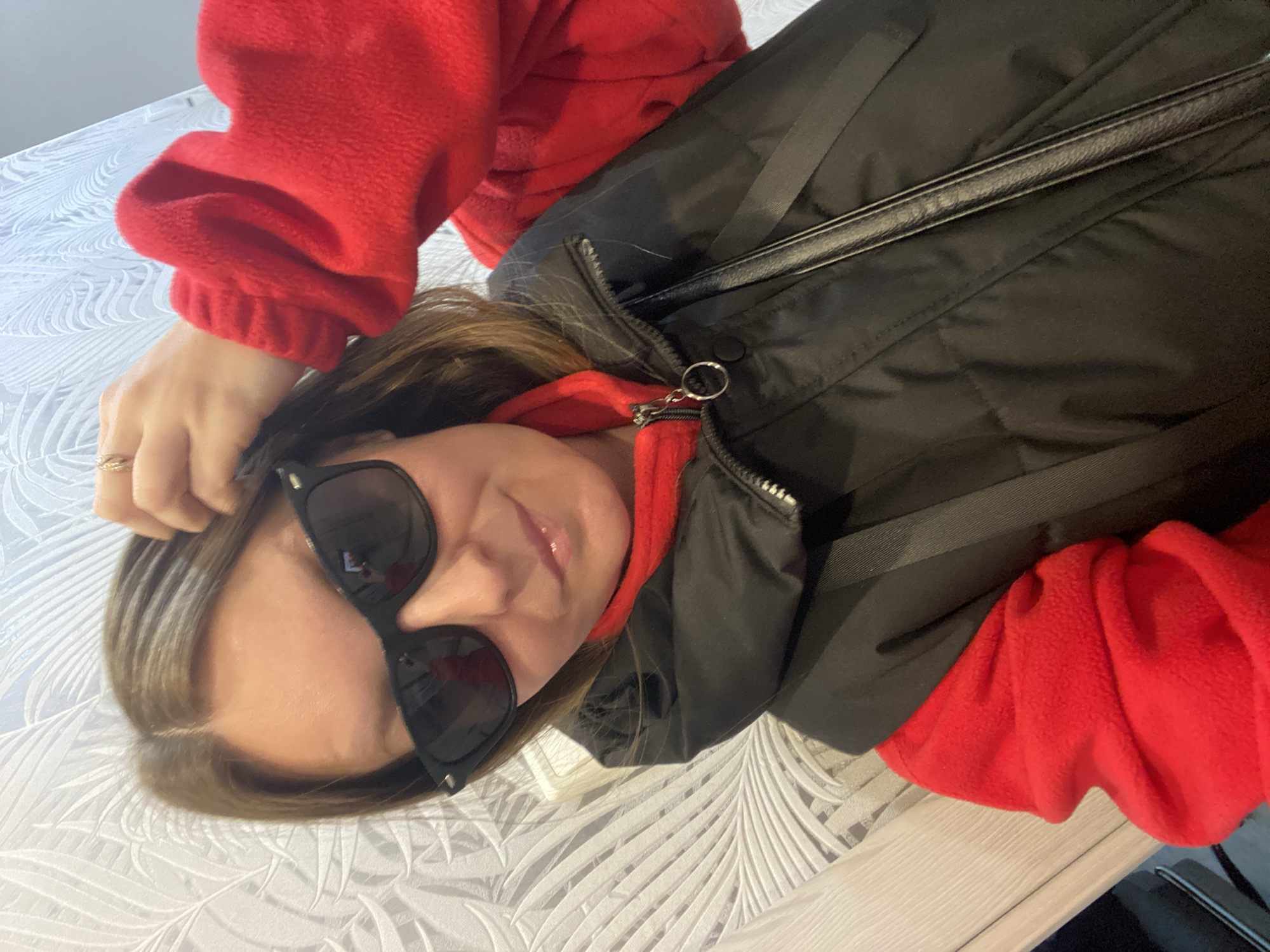 A woman is lying on a white surface with a leaf pattern. She wears a red fleece jacket and a black puffer vest. She is also wearing black sunglasses. Her right hand is touching her hair. The location is not provided in the image and cannot be determined.
