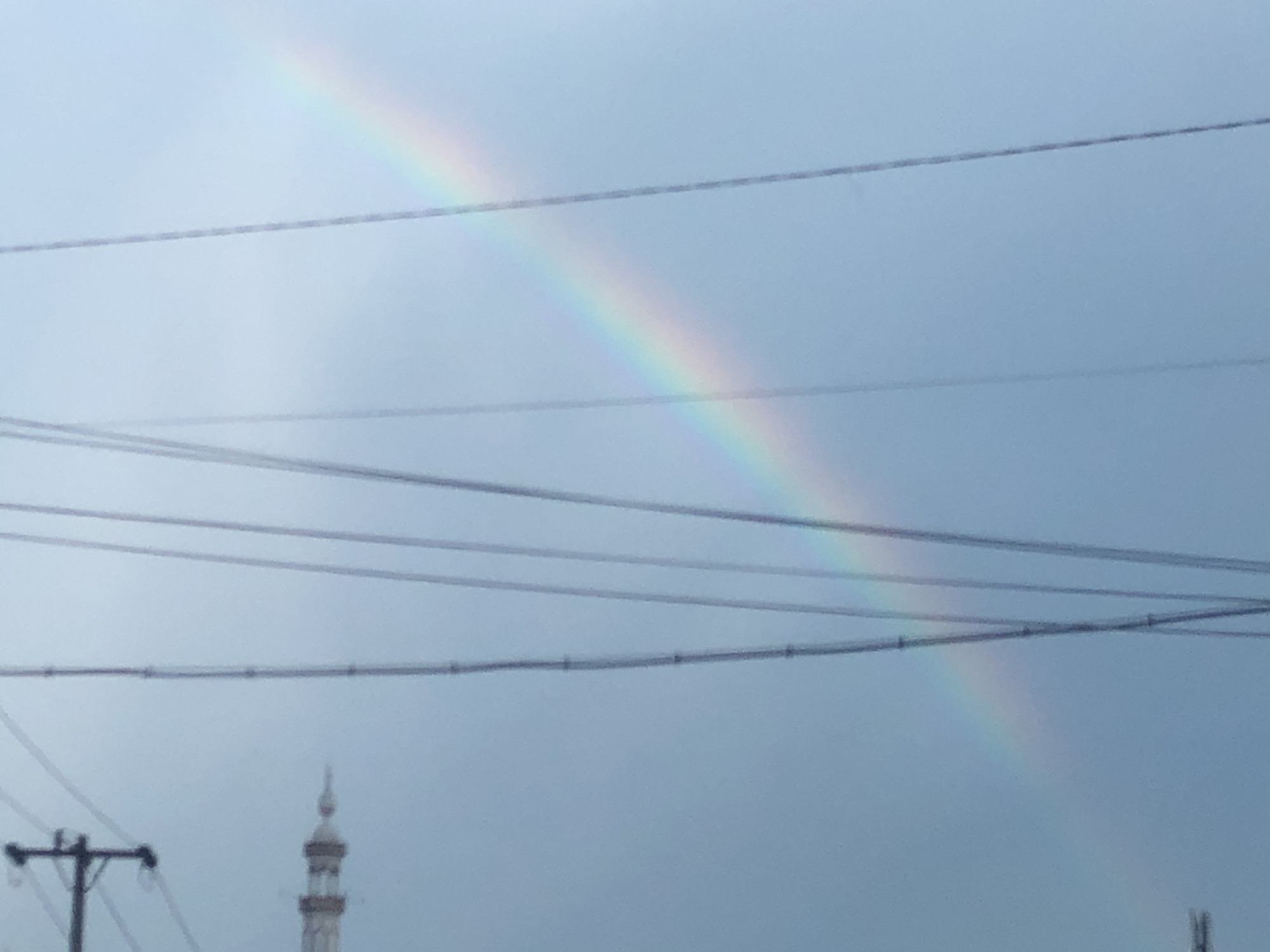 The image shows a segment of a rainbow in a sky of overcast conditions. Several telephone or electrical wires crisscross the lower portion of the rainbow. In the bottom left corner is a utility pole. To the left of the center, the minaret of a mosque is visible. The picture appears to be taken in an urban environment, likely in the City of Medan, Indonesia, as specified.