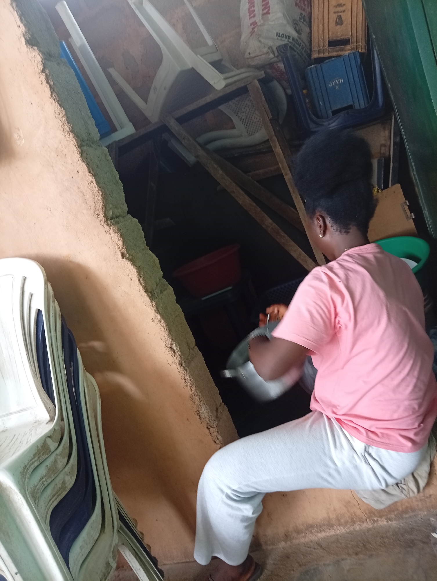 An African woman with an afro hairstyle is crouched in a cluttered storage area. She is wearing a pink t-shirt, grey pants, and brown shoes. She is holding a metal pot. The storage area contains stacked white plastic chairs, bags of flour, blue crates, wooden planks, and a red plastic basin. The location is likely Jos, Nigeria, based on the appearance of the scene and objects.