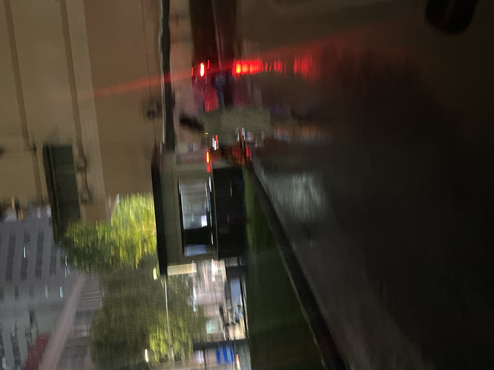 This blurry night image, captured in Yuzhong District, China, depicts an outdoor urban environment with wet, reflective ground. In the left background, indistinct tall buildings with horizontal light streaks are visible above blurred green trees, accompanied by a streetlamp. In the midground, a small, dark-windowed booth or building stands adjacent to a grassy patch. A highly blurred individual, wearing light-colored attire, is observed walking from left to right on a wet paved surface. To their right, a blurred red vehicle with illuminated taillights is present on the wet pavement. The wet ground prominently reflects the red taillights, creating distinct streaks of light in the foreground and midground. A larger building or structure with horizontal bands occupies the right background.