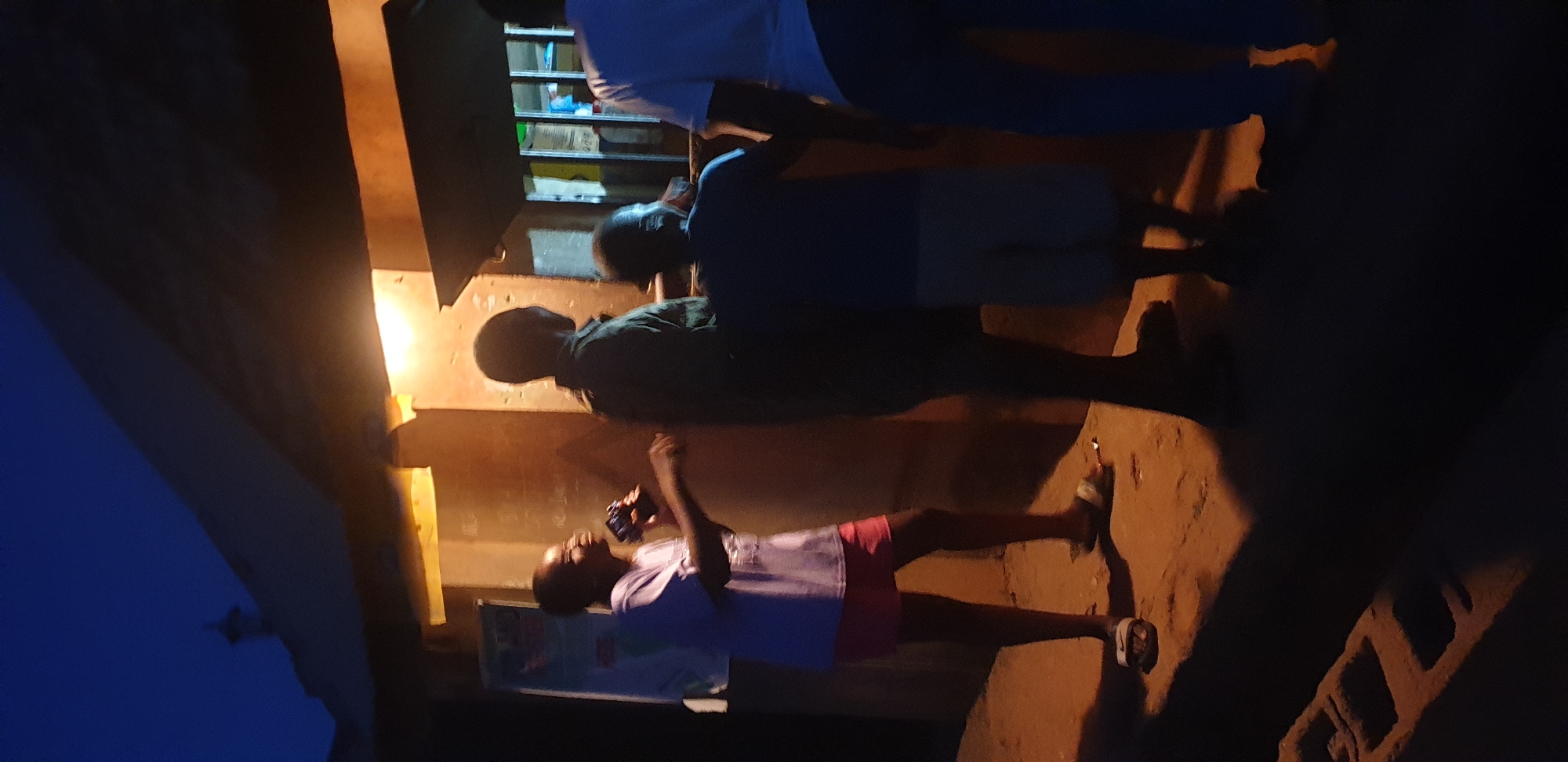The image, taken in Barden Yana, Nigeria, depicts a nighttime scene outside a small, rustic commercial stall. The stall, possibly constructed from concrete or mud brick, features a sloped roof and a service window or opening. A single, exposed artificial light source, likely a bare bulb, is positioned above this opening, casting a warm, yellow-orange illumination on the immediate area. A lighter-colored poster or sheet of paper is visible on the wall to the left of the opening.
Four individuals are gathered around the stall. From left to right:
1. A young person, possibly female, is standing in the foreground, wearing a light-colored t-shirt and pink shorts or a short skirt, with light-colored slide sandals on their feet. They are holding a dark-colored object, possibly a drink or a phone, up to their mouth or face, and appear to be smiling. Their body is slightly turned towards the stall.
2. A male individual stands facing the stall with his back mostly to the camera. He is wearing a dark, possibly long-sleeved, shirt and dark pants, largely silhouetted against the stall's light.
3. A child, identifiable by their smaller stature, is positioned directly in front of the stall's opening, facing it with their back to the camera. They are wearing a dark blue t-shirt and lighter-colored shorts.
4. Another male individual is positioned to the right of the child, also facing the stall with his back largely to the camera. He is wearing a light blue t-shirt and dark blue or black pants.
The ground beneath the individuals is unpaved, appearing to be dirt or sand, and reflects the warm glow of the artificial light. Shadows of the figures extend from their feet towards the lower right of the frame. In the lower left corner, a concrete block or similar construction material is partially visible in shadow. A small, light-colored object, potentially litter, lies on the ground near the feet of the leftmost person. The sky above is dark blue, indicating night, with no visible celestial bodies.