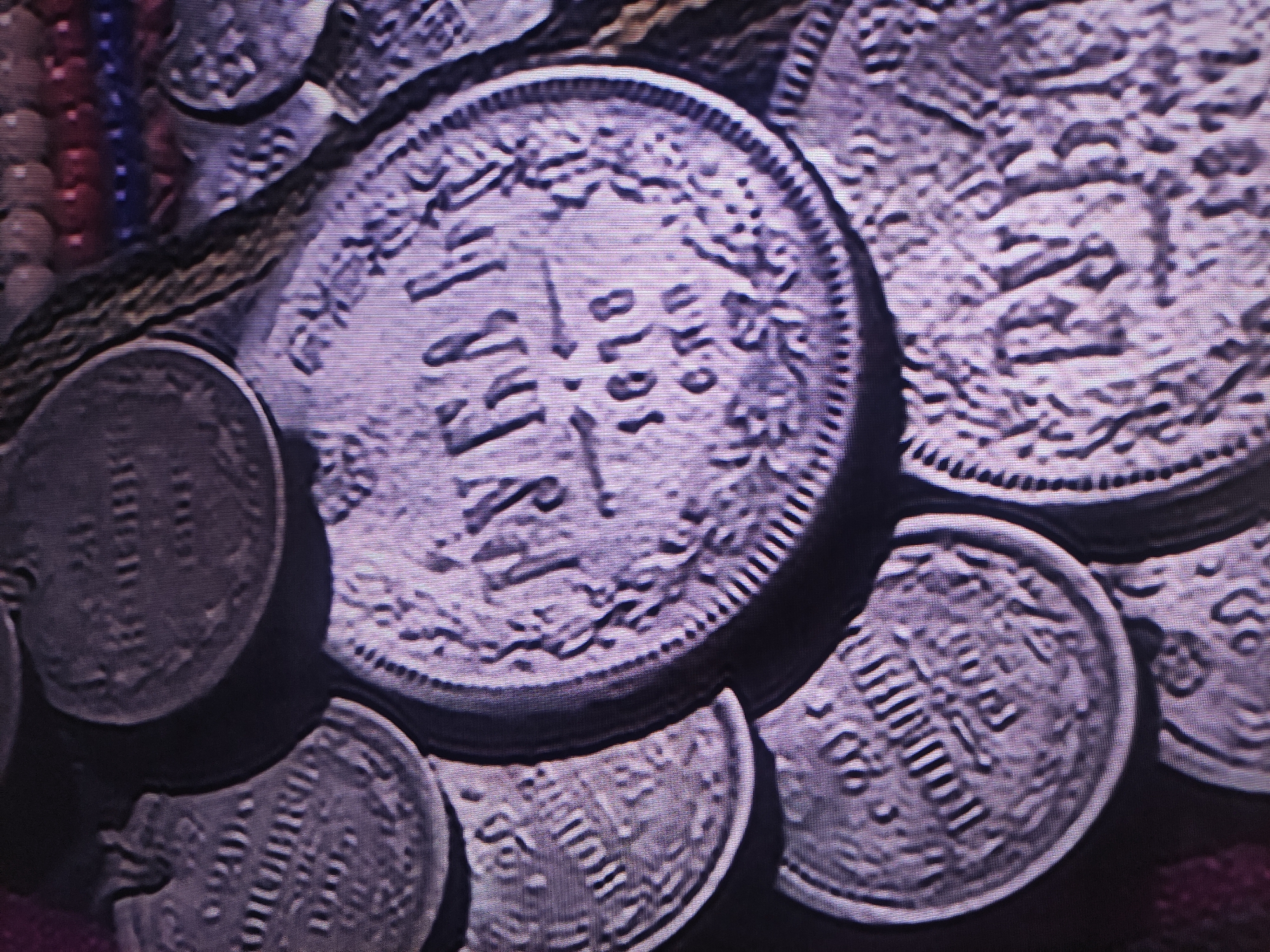 The image presents a close-up view of several old, metallic coins, possibly silver, laid out on a dark, textured surface. In the upper left corner, strands of colorful beads, including red and blue, are partially visible, suggesting they are part of a necklace or similar decorative item.
The most prominent coin, centrally located, is round with a distinct reeded edge. Its surface features intricate relief designs and clearly legible text, with the year "1910" prominently displayed in the center. Surrounding this date are characters that appear to be Cyrillic script, though their exact translation is obscured by the image quality and potential wear on the coin.
Other coins of varying sizes are scattered around the central piece, some partially overlapping. These also bear designs and less distinct text. One smaller coin in the lower right foreground shows numerical characters, potentially another date, but it is not fully clear. The coins generally exhibit a muted purplish-grey tone, characteristic of aged metal.
The image has a somewhat pixelated or low-resolution quality, with noticeable horizontal lines, suggesting it might be a photograph of a screen displaying the coins. The lighting emphasizes the raised details and textures of the coins. There are no people visible, and no specific activity is taking place beyond the apparent display of these historical artifacts. The provided context indicates a location in Pavlodar, Kazakhstan, but the image itself contains no specific visual cues to confirm this setting for the coins or their arrangement.