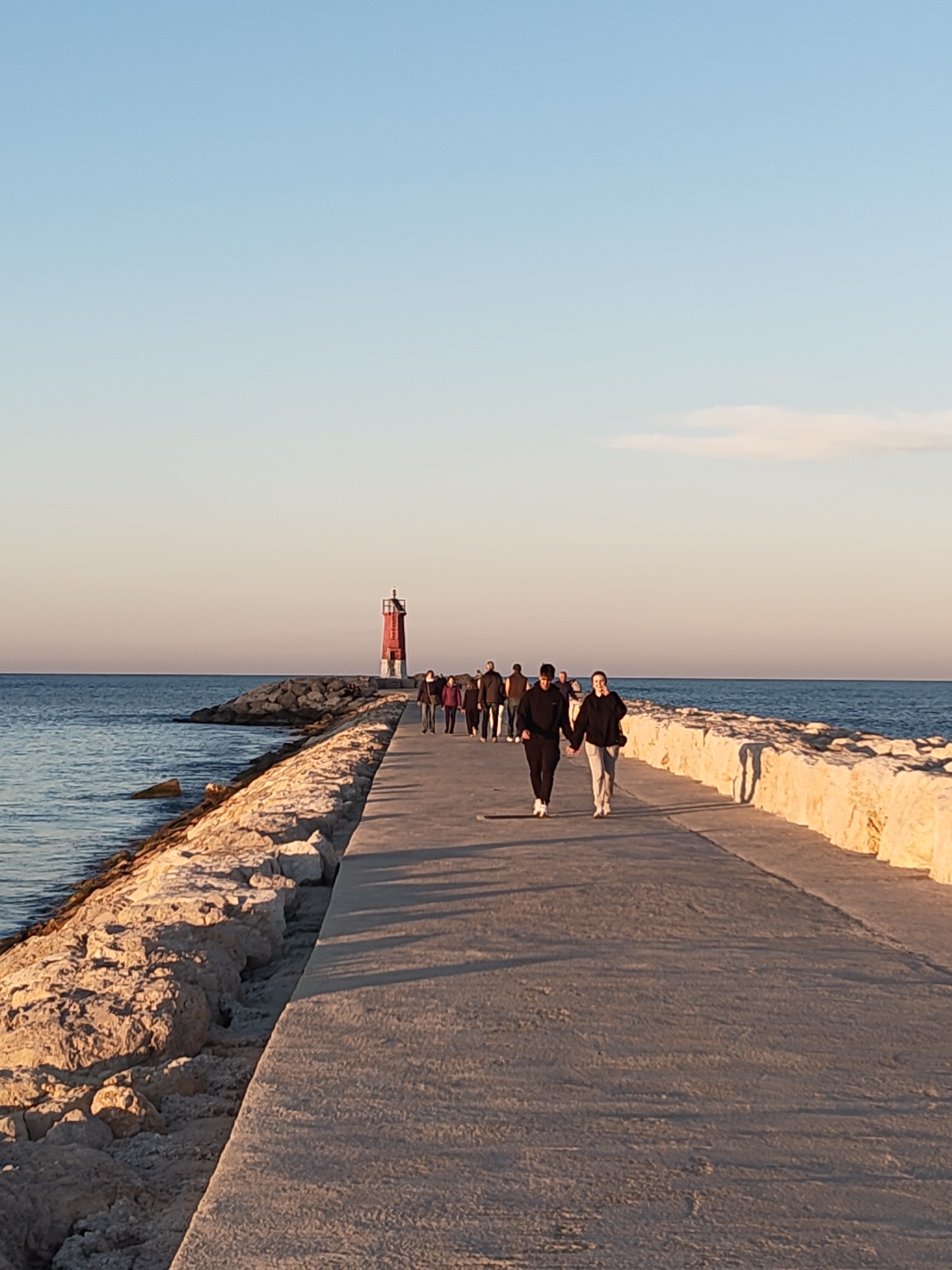 A long, concrete pier extends into the sea, with a line of people walking towards a red lighthouse at the end. The pier is lined with large, irregularly shaped white rocks on either side. The water is calm and blue-green. The sky is a gradient of light blue to pale orange, suggesting either sunrise or sunset. The two closest people hold hands. This is the Dénia pier in Dénia, Spain.