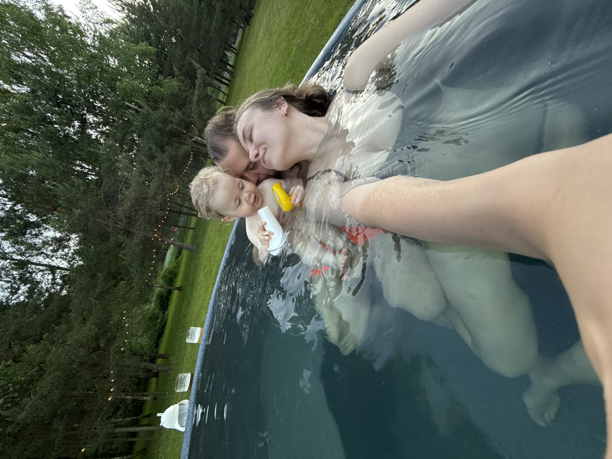 In Копнино, Russia, a family consisting of two adults and a young child is depicted within a portable, above-ground swimming pool. The woman on the right, with wet, light brown hair, is submerged up to her shoulders, wearing a dark-colored swimsuit. Her eyes are closed, and her head is tilted upwards and to the left, indicating a serene or relaxed expression. Her left arm is extended towards the camera, suggesting she is holding the device for a selfie. To her left, partially behind her, is a man with dark hair, looking down at the child with a gentle expression. Between the two adults is a young child with blonde, curly hair, holding a white plastic bottle in the left hand and a yellow plastic toy in the right hand, looking down at the objects. The child is wearing red swimwear bottoms visible underwater. The pool water is clear, revealing the submerged limbs of the individuals. On the blue-grey rim of the pool, to the far left, rests a white, inverted object resembling a hat or cover, alongside two glasses containing a light amber liquid with foam, suggestive of beverages. The background features a vibrant green lawn leading to a dense line of various trees, including conifers and deciduous types. A string of small, warm-toned lights is suspended horizontally between the trees, creating an evening or twilight ambiance.
