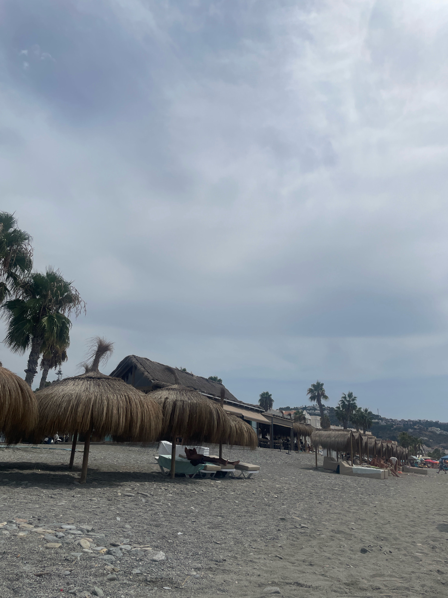 The image depicts a cloudy day on a beach in La Herradura, Spain. In the foreground, a pebble-strewn sandy beach is visible with several thatched umbrellas providing shade. Underneath one umbrella, a person lies on a lounge chair. In the background, there are more thatched umbrellas, lounge chairs with people relaxing, and a building with a thatched roof that appears to be a beachside restaurant or bar. Palm trees are scattered throughout the scene, with a hill covered in buildings and vegetation in the distance. The overall atmosphere is relaxed and casual, typical of a beach resort.