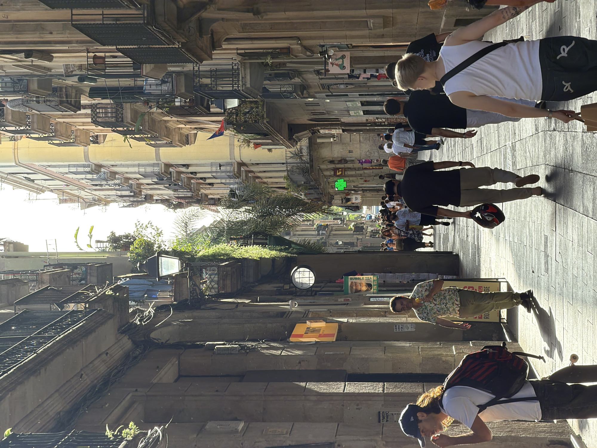 The image depicts a sunlit narrow street in Barcelona, Spain, likely in the Gothic Quarter, characterized by tall, old buildings with balconies and an overhead pedestrian walkway. Sunlight casts strong shadows on the cobblestone pavement. Several people are walking in both directions along the street, some appearing to be tourists. On the left side, a young man with a baseball cap and a backpack is visible, while further down, another man in a patterned shirt is eating an ice cream cone. On the right side, a group of people are walking away from the camera, including a person in a white tank top and jeans carrying a bag. Storefronts and signage are visible on the building walls, including a green neon sign and a sign with a scooter graphic. The architecture features weathered stone and stucco facades, and ornate iron railings on the balconies. Vegetation, including palm trees and ivy, is integrated into the urban environment.