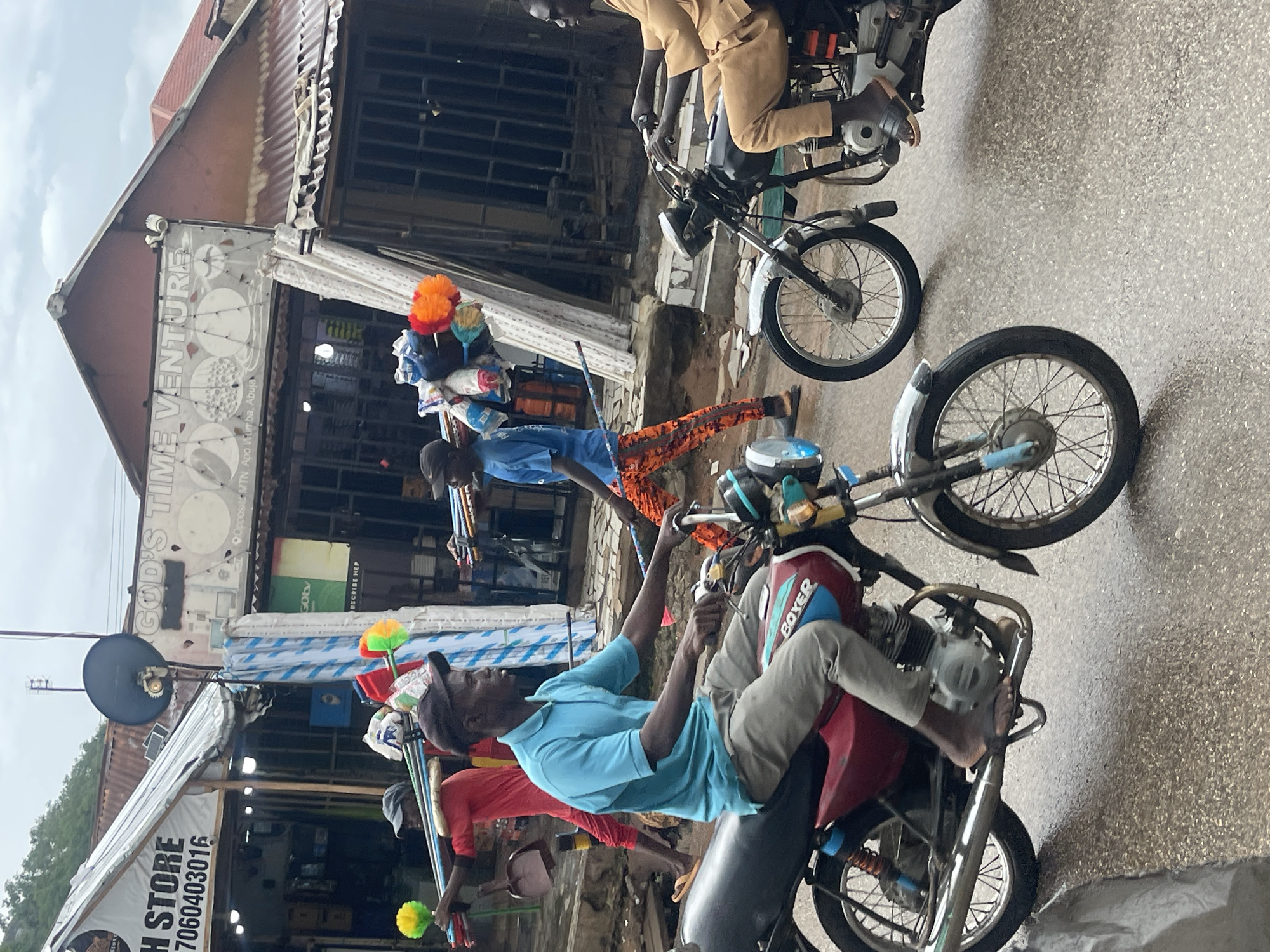 This image captures a bustling street scene in Abuja, Nigeria, featuring multiple individuals, motorcycles, and storefronts. The sky appears overcast, and the road surface looks damp, suggesting recent rain or a humid day.
In the foreground and midground, several men are present. One man, wearing a light blue polo shirt, grey trousers, and a dark cap, is seated on a red "BOXER" brand motorcycle, holding the handlebars. To his right, another man in a red shirt, dark pants, and a cap is pushing a red motorcycle, carrying multiple colorful whisk-like items, likely for sale, on a stick over his shoulder. Further back and slightly to the left, a third man, dressed in a blue shirt and orange patterned trousers, is walking or standing, also carrying a bundle of items, including similar colorful whisks, on his head or shoulder. In the background, another man in a tan outfit is seated on a beige motorcycle.
The scene is set against a backdrop of local shops with corrugated iron roofs. Signs are visible on the shopfronts. One prominent sign reads "GOD'S TIME VENTURE" and "Opposite MTN, Apo Wurrba, Abuja," confirming the location. Another sign, partially visible, reads "H STORE" followed by a phone number "7060403016." Other visible text includes "GOtv" and "OSCRIBE HEP" (possibly a partial "SUBSCRIBE HERE"). A satellite dish is also mounted on one of the buildings. The people in the image appear focused on their activities, conveying a sense of routine daily life in a lively urban environment.
