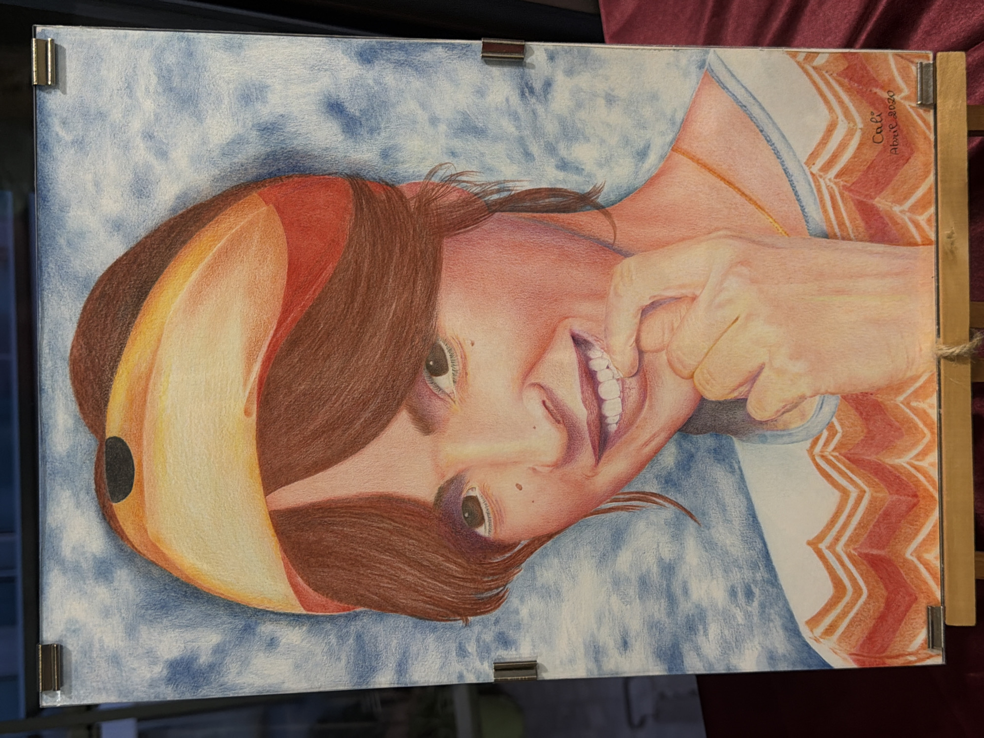 A colored pencil drawing on paper depicts a young person with reddish-brown hair styled with a yellow and orange headband. The individual has a bright smile, with their index finger touching their lower lip. They are wearing a white shirt with a blue neckline and an orange and white chevron pattern around the hem. The background is a mottled blue. A signature and date, "Cali Abril 2020," are visible in the lower right corner. The artwork is framed and displayed on an easel. The specific location in Elche/Elx, Spain, is not ascertainable from the image.