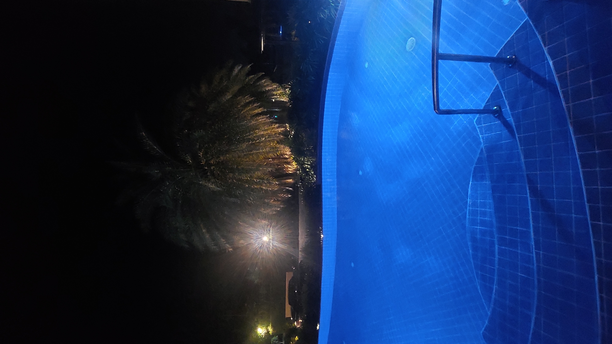 The image shows a nighttime scene in Baan Mae Nam, Thailand. On the right is a brightly lit swimming pool with a metal ladder leading into the water. The pool has tiled steps leading down into it. To the left of the pool is a dark area with a palm tree partially illuminated by a ground-level light. A taller building is visible in the background, also lit from below.