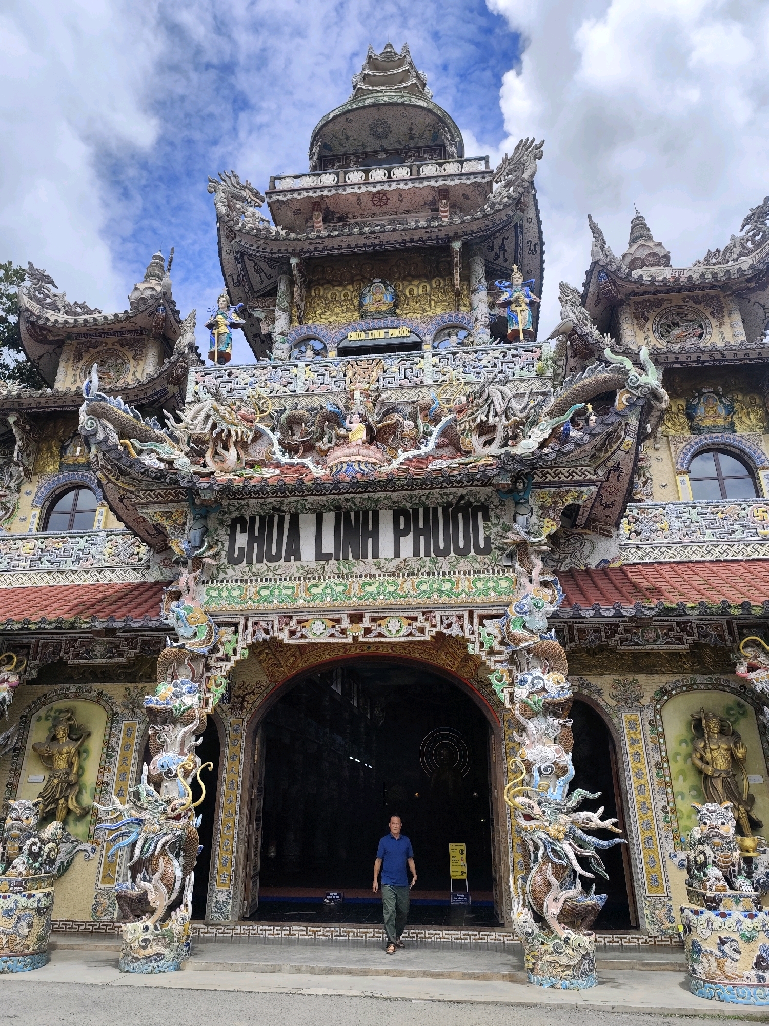 A man walks towards the entrance of the Linh Phuoc Pagoda in Xuân Thành, Vietnam. The pagoda is an ornate Buddhist temple constructed primarily from broken ceramic and glass. The facade is adorned with intricate mosaics depicting dragons, mythical creatures, and Buddhist figures. The entrance is framed by two large, colorful dragon sculptures on pillars. The sky above is partly cloudy. The man is wearing a blue polo shirt and green pants. He is walking on a paved walkway leading to the temple.