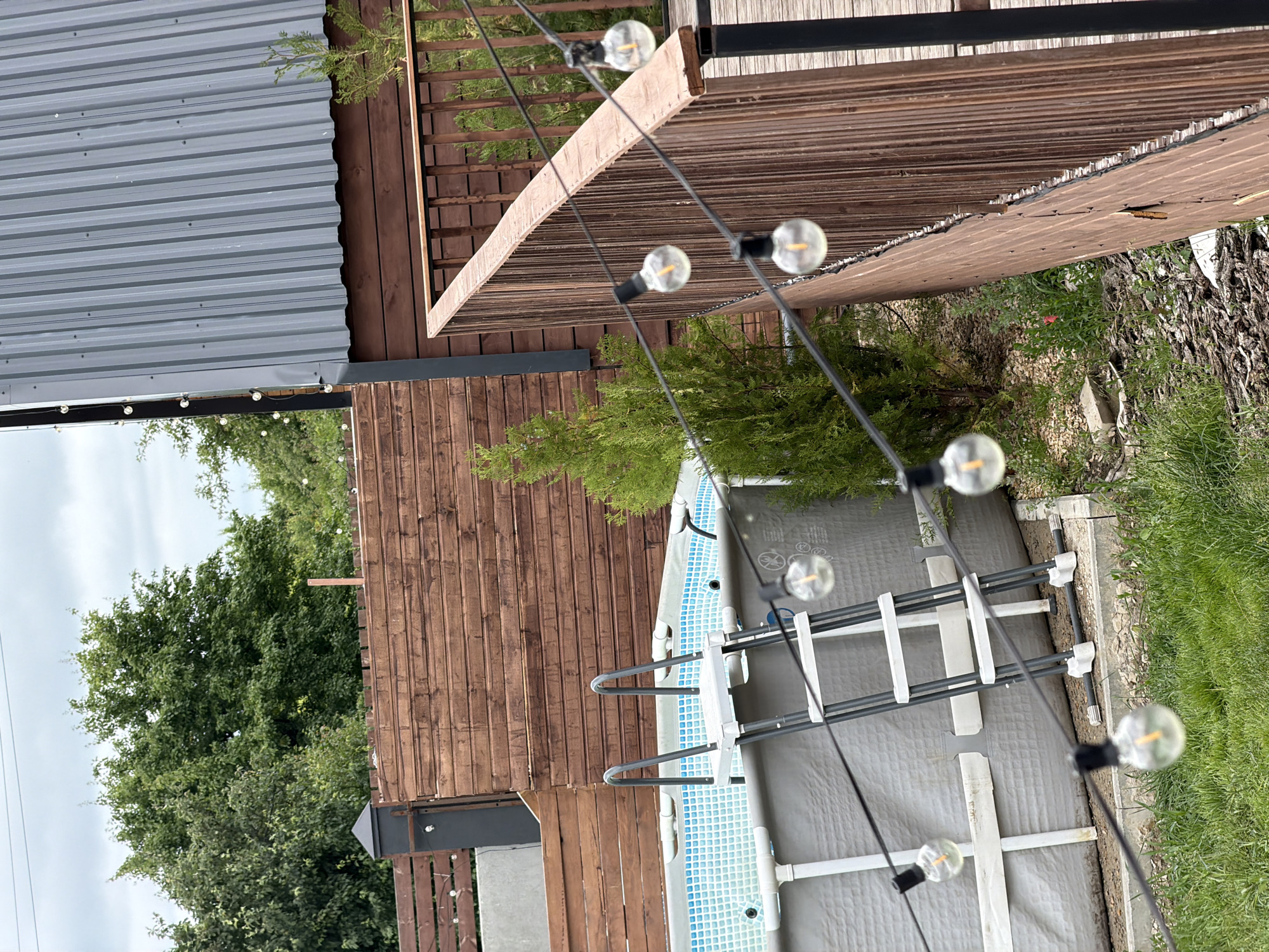 The image depicts an outdoor scene in a residential yard, likely in the city of Семилуки, Russia, featuring a backyard setup with a swimming pool and a wooden deck. A grey, above-ground swimming pool with a blue tiled interior and a ladder is visible on the left side of the frame. String lights with clear Edison-style bulbs hang across the scene, some in focus and others blurred. A wooden fence and a section of a wooden deck with vertical railings occupy the right and central portions of the image. Beyond the immediate yard, lush green trees and a cloudy sky are visible. The overall impression is of a leisure space prepared for outdoor activities.