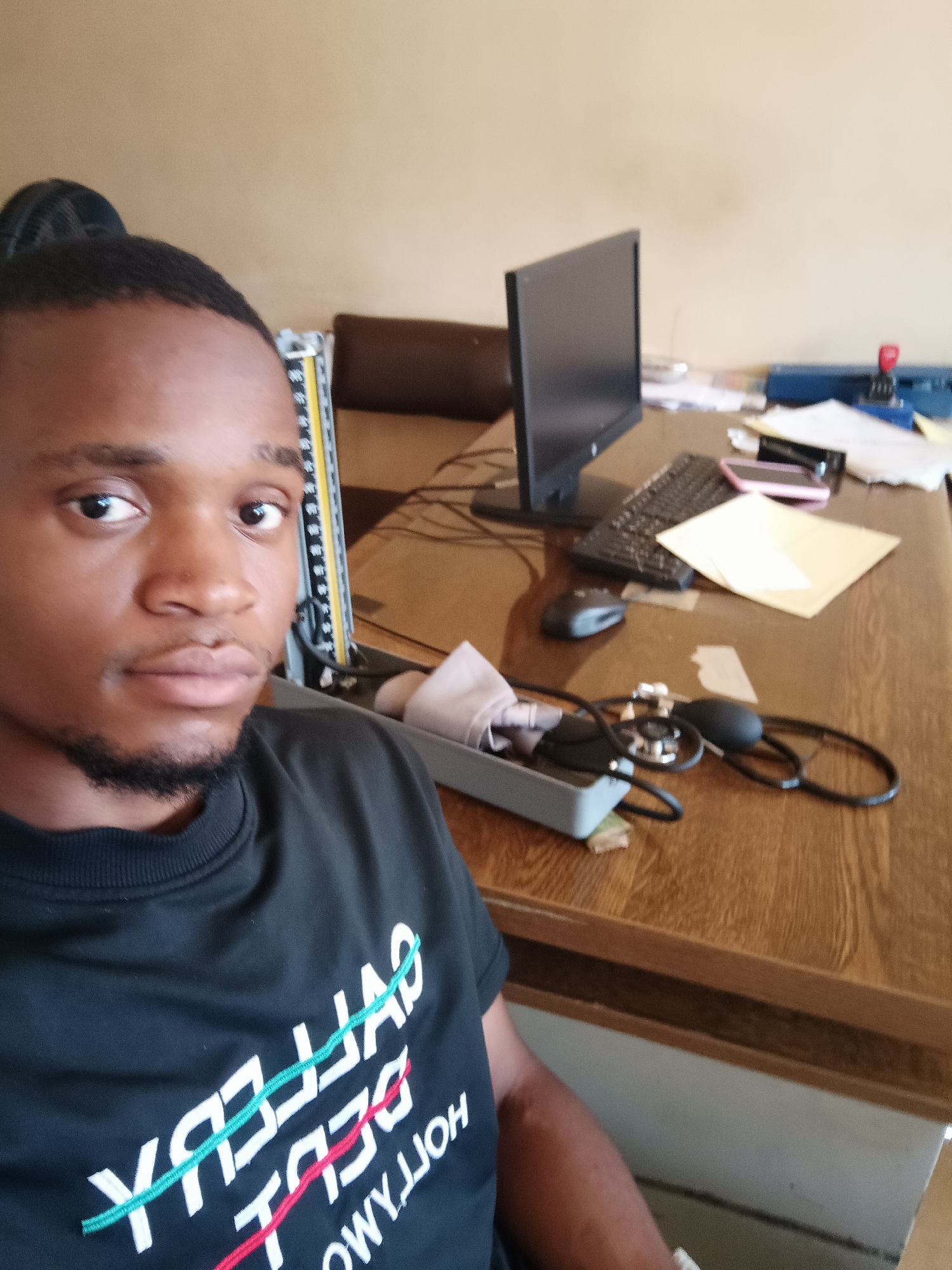 A young Black man is featured in a close-up selfie, looking directly at the camera with a neutral expression. He has short dark hair and a neatly trimmed beard or goatee. He is wearing a black crew-neck sweatshirt with prominent mirrored text on the front.
The setting appears to be an indoor office or clinic, with the man seated at a wooden desk. On the desk, various items are scattered, suggesting a workspace. These include a black computer monitor, a keyboard, a grey computer mouse, and several papers or documents, one of which is a light yellow folder or envelope. A pink smartphone is also visible near the keyboard.
More notably, medical equipment is present on the desk: a blood pressure monitor (sphygmomanometer) with its cuff, bulb, and stethoscope-like tubing, and a tall, yellow-and-black striped device that resembles a measuring tape or part of a medical scale. A dark blue stapler is also visible in the background. The walls are a light, neutral color.
The visible text on the man's black sweatshirt is mirrored, but can be deciphered as:
- Top line: "GALLERY" (with a teal stripe running through it)
- Middle line: "DEBUT" (with a red stripe running through it)
- Bottom line: "HOLLYWOOD"
The general lighting suggests it is daytime. The scene provides location cues indicating a professional or medical environment within Kakuri Gwari, Nigeria.