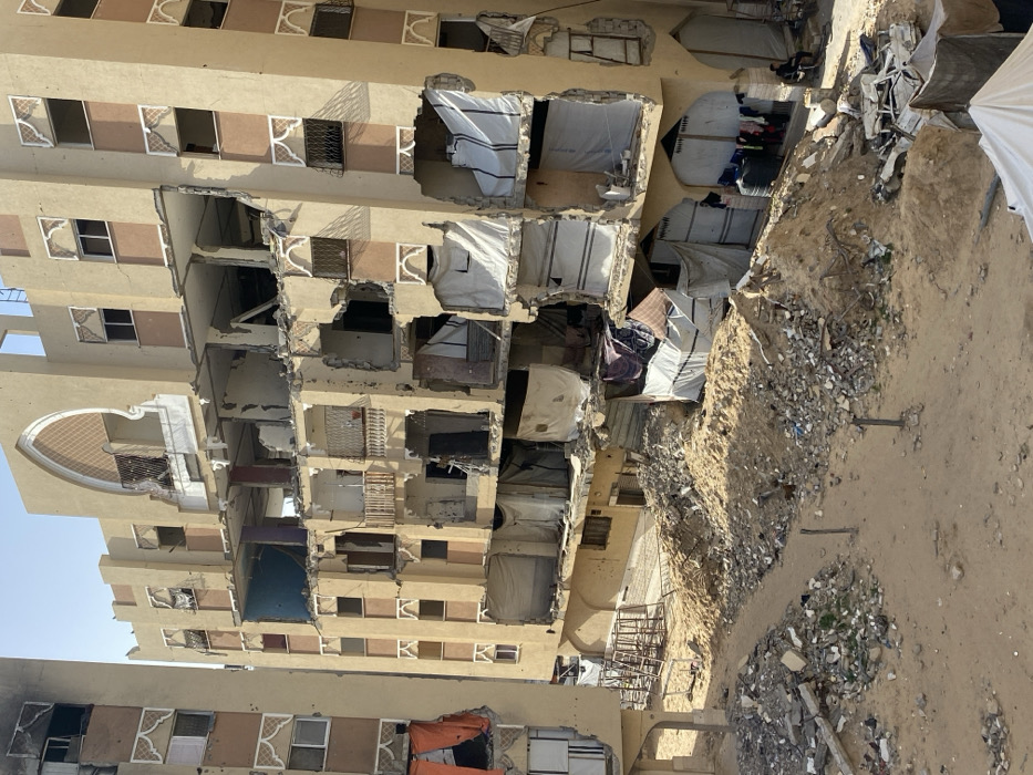 An elevated view captures a desolate urban scene in Khan Yunis, Palestinian Territory, dominated by two severely damaged multi-story residential buildings under a clear blue sky. The prominent building on the right, a tan-colored structure with ornate arched window details and balconies, exhibits extensive structural failure across at least six visible floors. Most exterior walls have been destroyed or collapsed, exposing interior rooms. Many of these exposed sections are covered with white, gray, or striped tarpaulins, acting as makeshift screens or enclosures. Concrete debris, twisted rebar, and shattered remnants of fixtures are visible within the open cavities. The building on the left, partially visible, shows similar severe damage, including blackened sections indicative of fire damage on its upper floors and orange tarpaulins covering lower exposed areas.
The ground between and in front of the buildings is a landscape of destruction, covered by large mounds of sand, dirt, and construction rubble, including concrete fragments, shattered glass, and assorted debris. A narrow, sandy path winds through the foreground rubble. In the lower right corner, near the base of the main building, a black water tank is visible next to a clothesline with various garments drying. A single individual is seated on a small chair or stool in this area, observing the scene. To the far left, indistinct metal structures and additional debris are present. The overall environment conveys widespread devastation and makeshift adaptation.