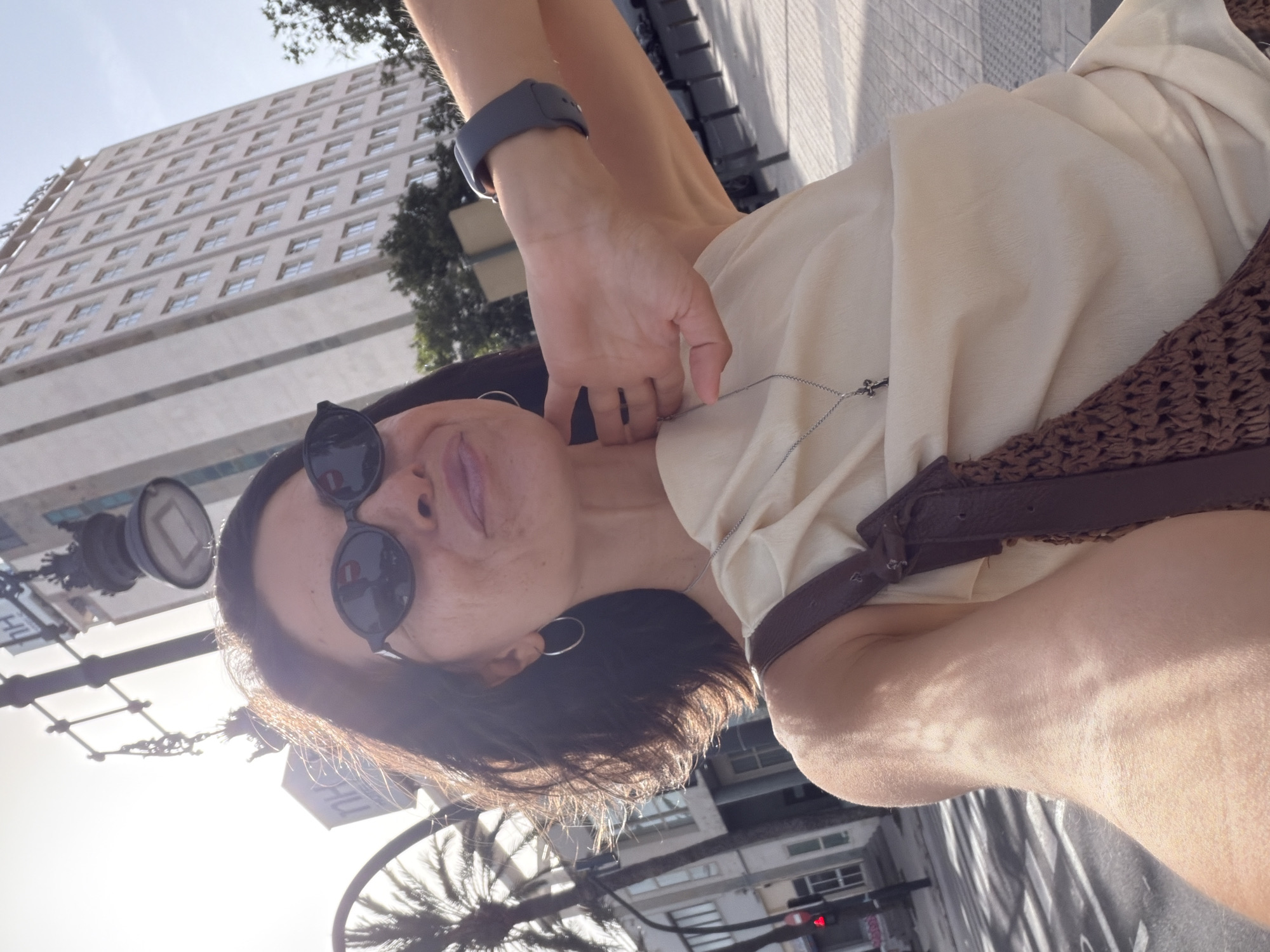 A woman, viewed from the chest up, is taking a selfie outdoors in front of a tall, light-colored building. She is wearing dark oval sunglasses, a light-colored sleeveless top, and a necklace with a small cross pendant. A brown woven bag with leather straps is slung over her left shoulder. Her right hand is resting on her neck. The background shows a palm tree on the left and a street scene with a traffic light. The sun is shining brightly, casting shadows.