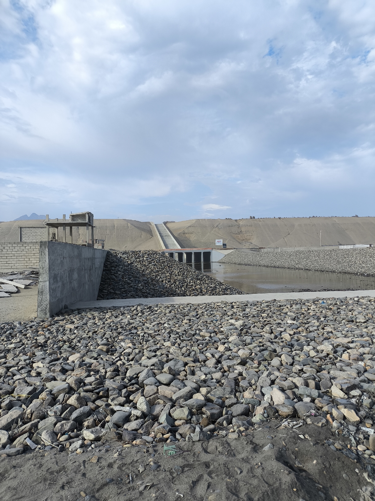 The image depicts a landscape featuring a dam or flood control structure, located in an area with sandy terrain. In the foreground, a field of large, gray rocks dominates the view, leading up to a concrete wall and what appears to be the start of a building construction. Behind this immediate foreground, a body of water extends towards a dam-like structure with multiple openings. The dam is connected to a sandy, elevated area that appears to have steps leading up its side. The background consists of a light blue sky with scattered clouds. No people are visible in the image. The city and specific location are unknown.