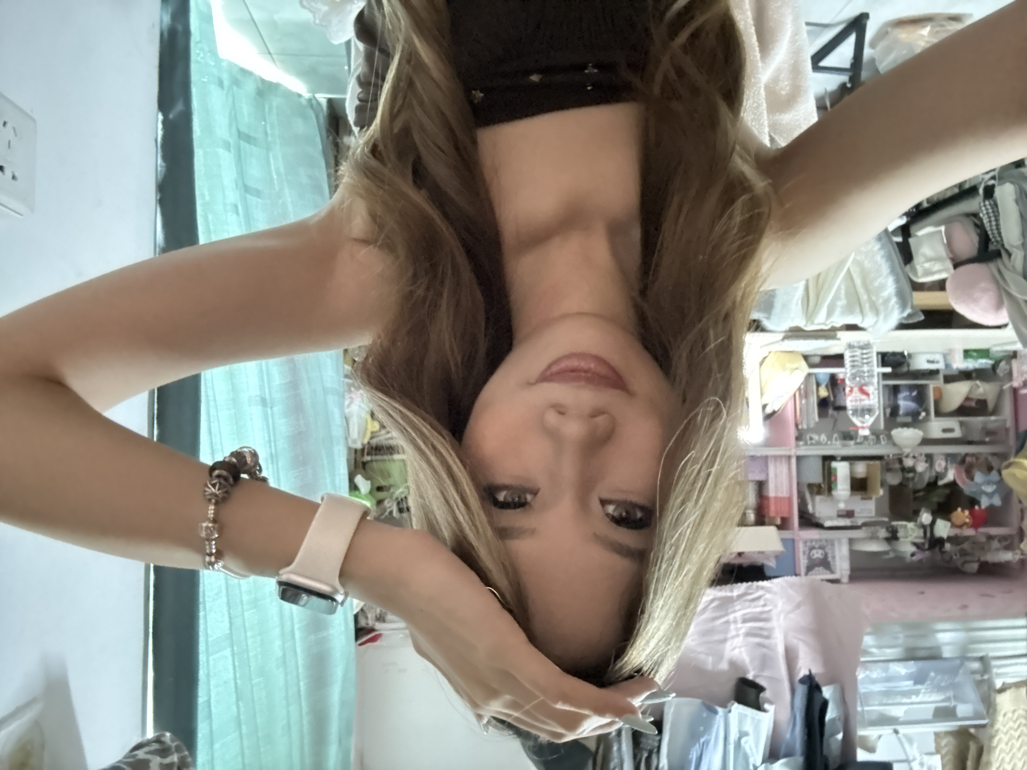 A woman with blond hair is upside-down in an indoor environment. She is wearing a black top and has jewelry on her left wrist, including a smartwatch. The background is cluttered with various objects on shelves and other items. The location in the city of Nan'an District, China cannot be determined from this image.
