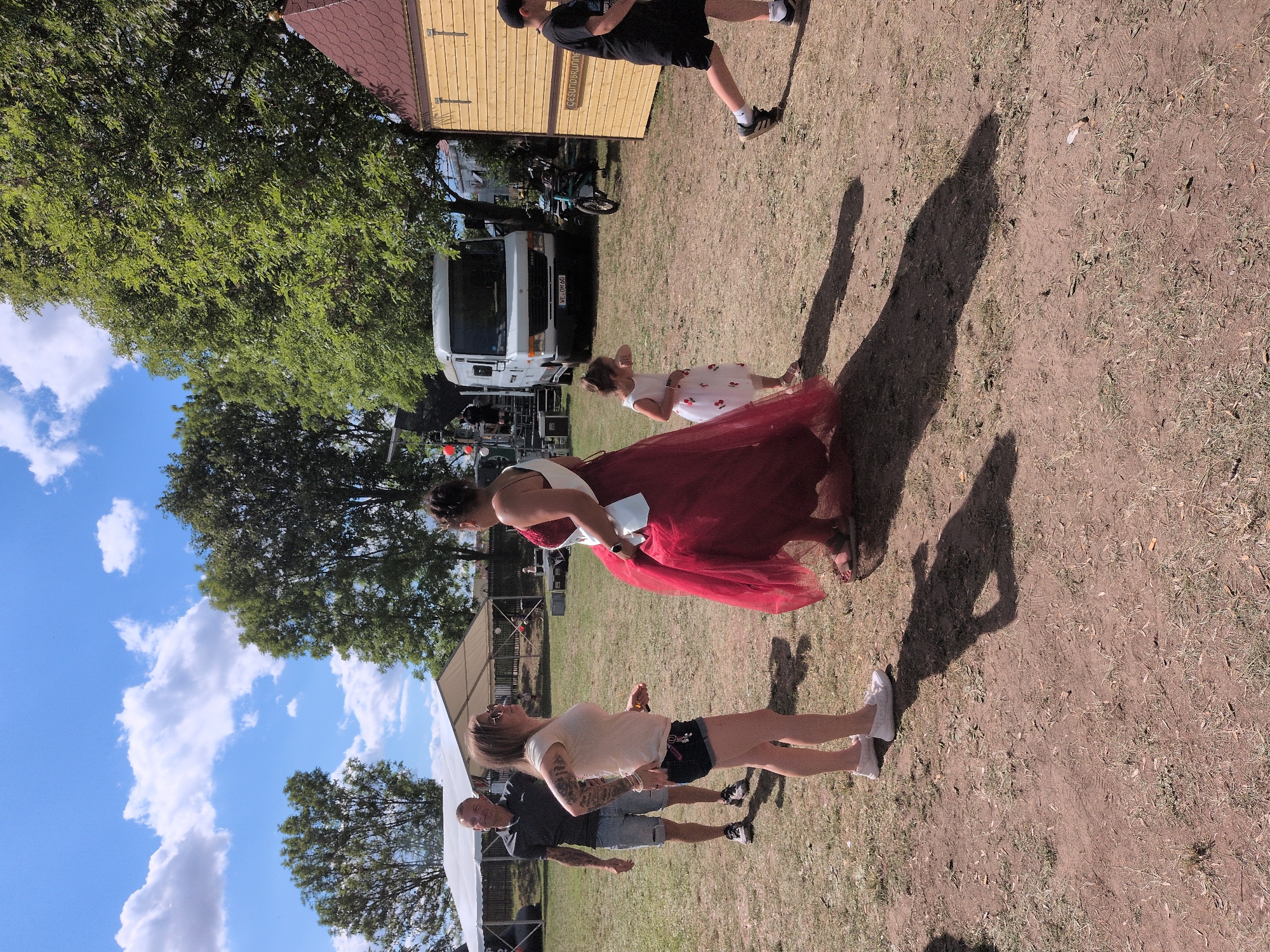 This image depicts an outdoor gathering in Rastenberg, Germany, possibly a fair or festival. The foreground is dominated by dry, dusty ground with sparse grass. In the center, a woman in a long, flowing red dress with a white sash is partially visible, appearing to be the focus of attention. To her left, a woman in a light-colored t-shirt and denim shorts, wearing sunglasses, stands with her back to the camera, her arm extended towards the woman in red. Behind them, a man in a dark polo shirt and denim shorts stands facing the group. To the right of the woman in red, a young girl in a white dress with cherry patterns is looking down and touching her face. Further to the right, a young boy in a black t-shirt and shorts walks away from the camera. In the background, a white van with a license plate reading "WE QM 60" is parked. Several trees with green foliage are visible, and a white tent structure is set up to the left. A small wooden building with a red roof and brown siding is situated on the right side of the image, with text that appears to read "Gesundbrunnen". The sky is blue with scattered white clouds.