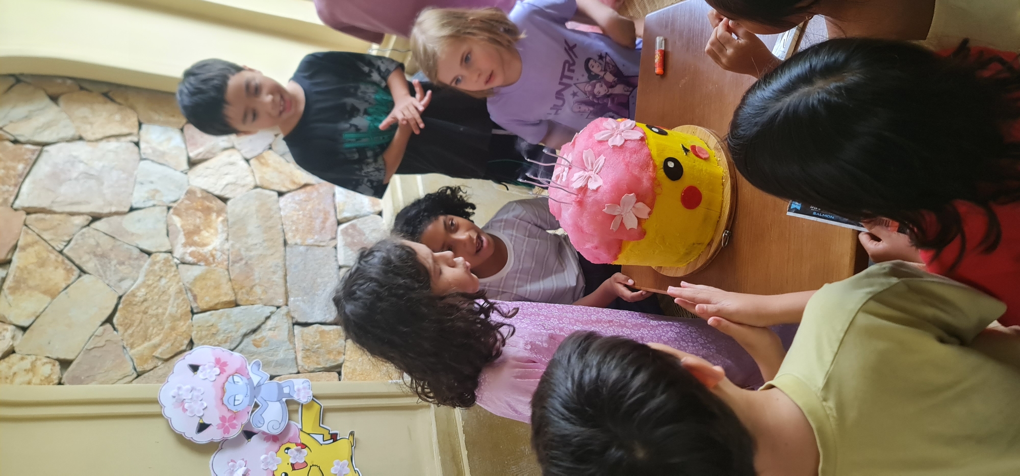 A group of children are gathered around a table with a Pikachu-themed cake, which is half yellow and half pink with flower decorations. The children are of various ethnicities and are wearing casual clothing. Some children are looking at the cake, while others are looking at each other. There is also a stone wall in the background and decorations of Pokémon characters on the wall. The location is Đà Nẵng, Vietnam.