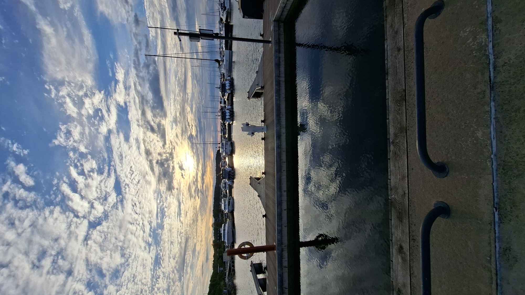 The image depicts a marina at sunset in Tallinn, Estonia. In the foreground, a concrete pier with wooden decking and metal railings overlooks calm water reflecting the sky and surrounding boats. Moored boats, including sailboats and motorboats, are visible in the mid-ground. The sky is filled with dramatic, textured clouds illuminated by the setting sun, which is partially obscured. The overall scene conveys a peaceful, end-of-day atmosphere in a coastal urban setting.