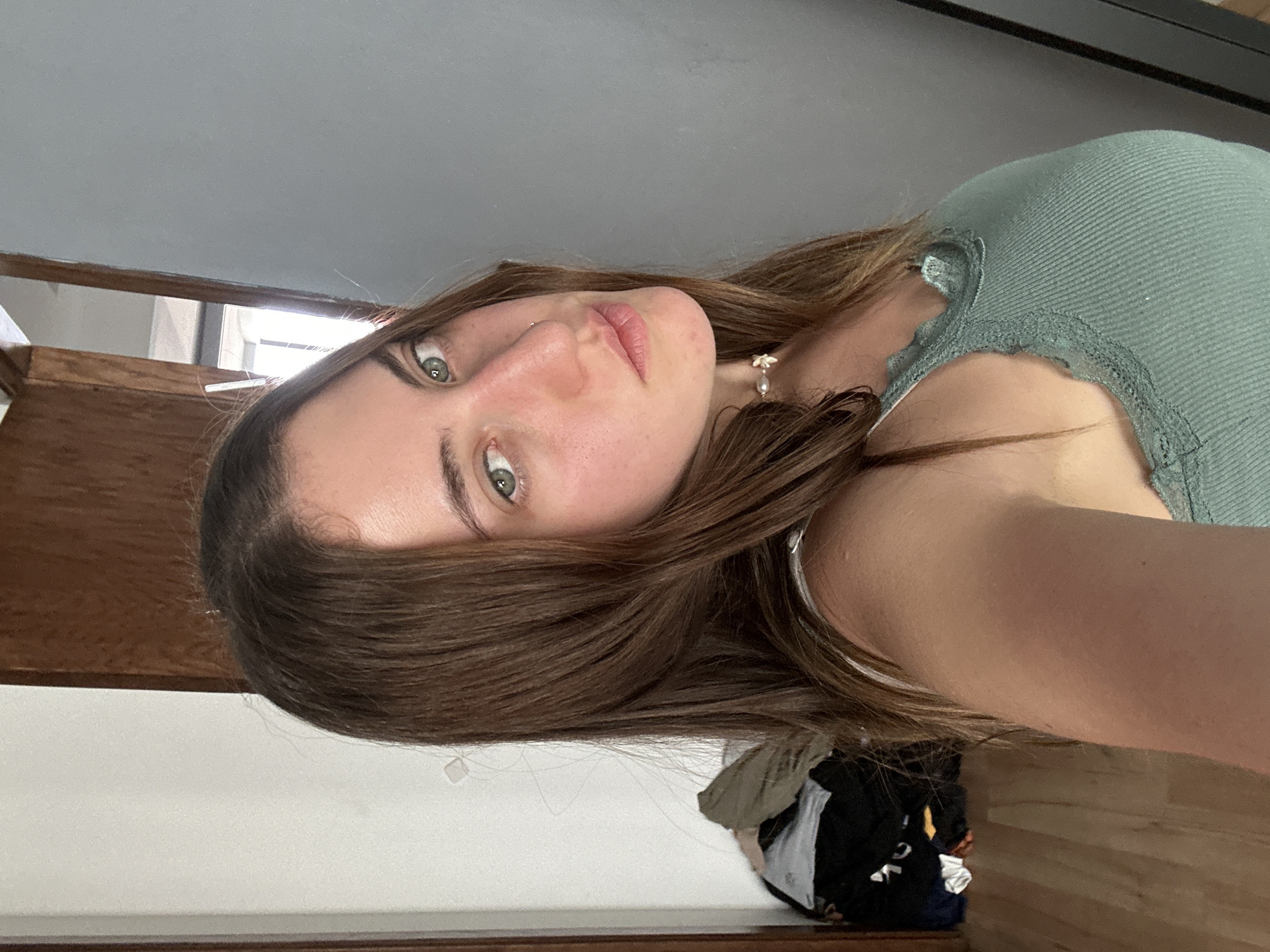 A young woman with brown hair and green eyes is centered in the image. She is wearing a light green, ribbed tank top with lace trim. She appears to be indoors, near stairs and a window. Clothes are piled on the floor. It is impossible to determine the specific location in Huixquilucan de Degollado, Mexico from this image.