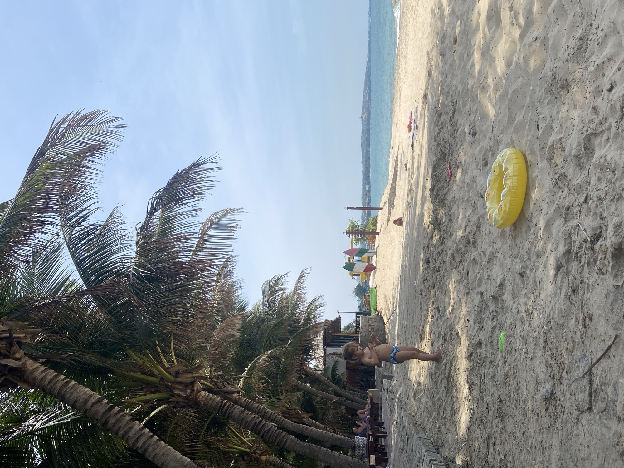 The image depicts a tropical beach scene in Phu Thuy Ward, Vietnam, under a clear blue sky. Tall palm trees line the left side of the frame, their fronds casting shadows on the sandy ground. In the foreground, a young child wearing blue and white patterned swim trunks stands on the sand, holding a stick. To the right of the child, a yellow inflatable ring lies on the beach. Further back, a few people are visible sitting at tables under a thatched-roof structure, and another individual is lying on a towel near the water. In the distance, the calm blue ocean meets the horizon, with a coastal town or resort visible on the far shore. The beach appears to be part of a resort or public recreation area, with surfboards and a yellow structure that could be part of a playground visible further inland. The overall atmosphere is one of leisure and relaxation.