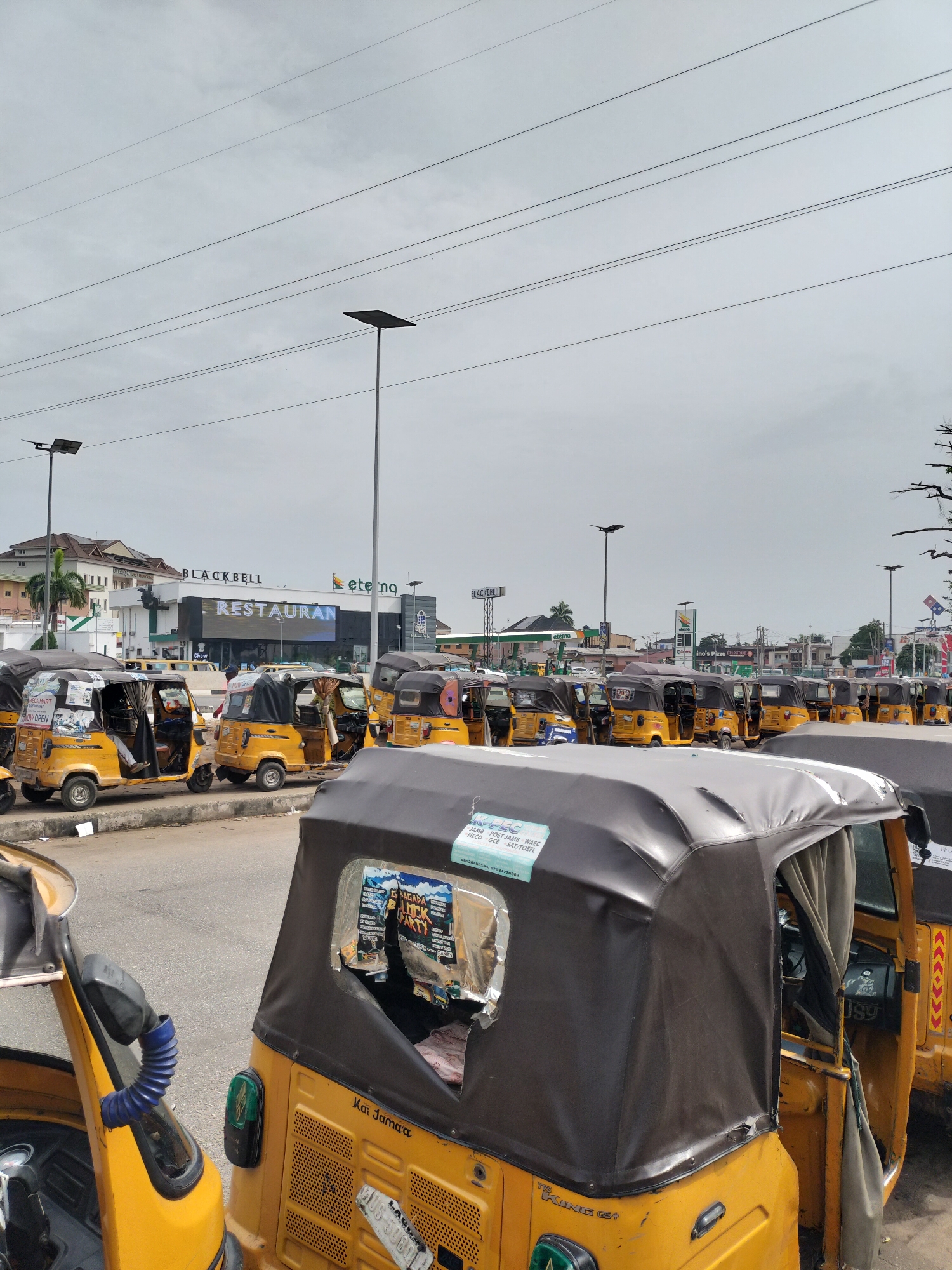 The image shows a line of yellow commercial tricycles (locally called "Keke") parked in an urban area. The setting is Lagos, Nigeria, evidenced by the Lagos State vehicle registration plates on the Keke. A multi-story building is visible in the background with signs that read "BLACKBELL" and "RESTAURANT." Overhead power lines and tall streetlights stretch across the sky. There are advertisements on some of the Keke. The sky is overcast. People are visible inside some of the vehicles.